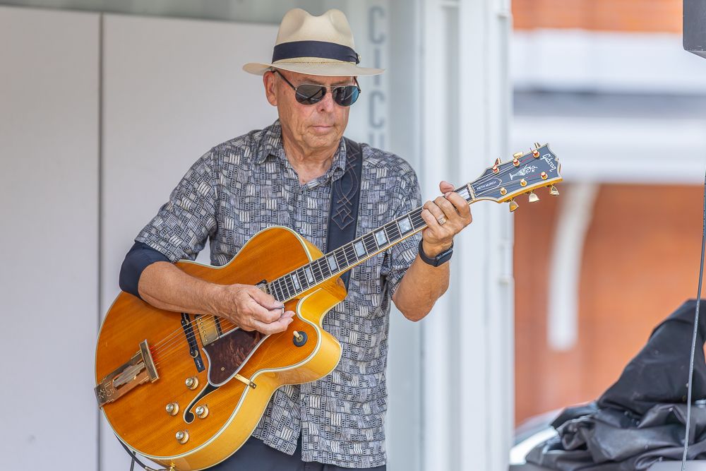 Musician plays an acoustic guitar in front of a bright window while wearing a floral shirt and sunglasses.