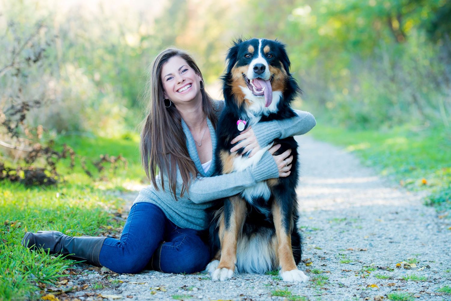 A Bernese Mountain Dog and companion sitting together on a nature path surrounded by green foliage.