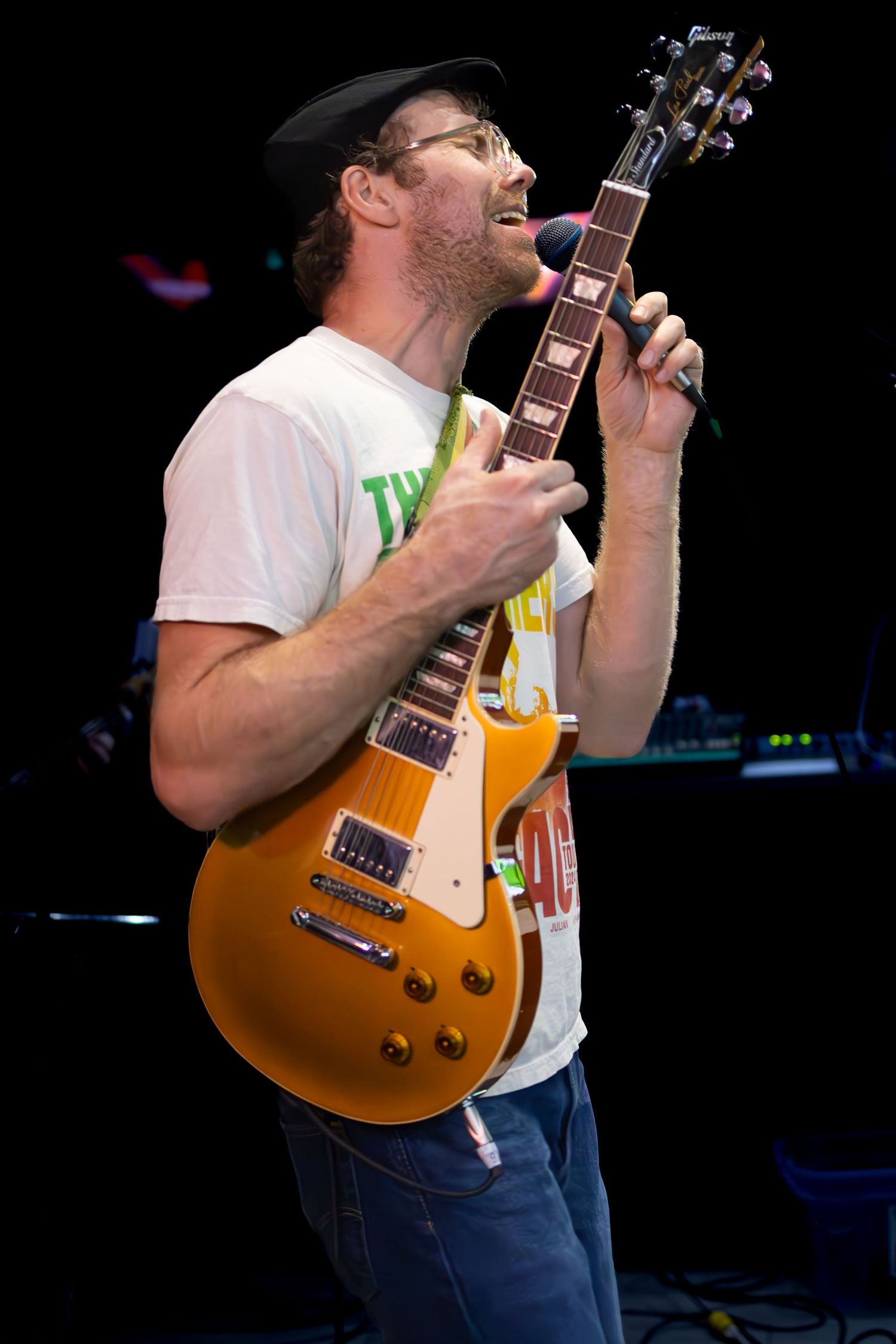 Musician performs on stage with a sunburst Gibson Les Paul guitar while wearing glasses, a white t-shirt and baseball cap.