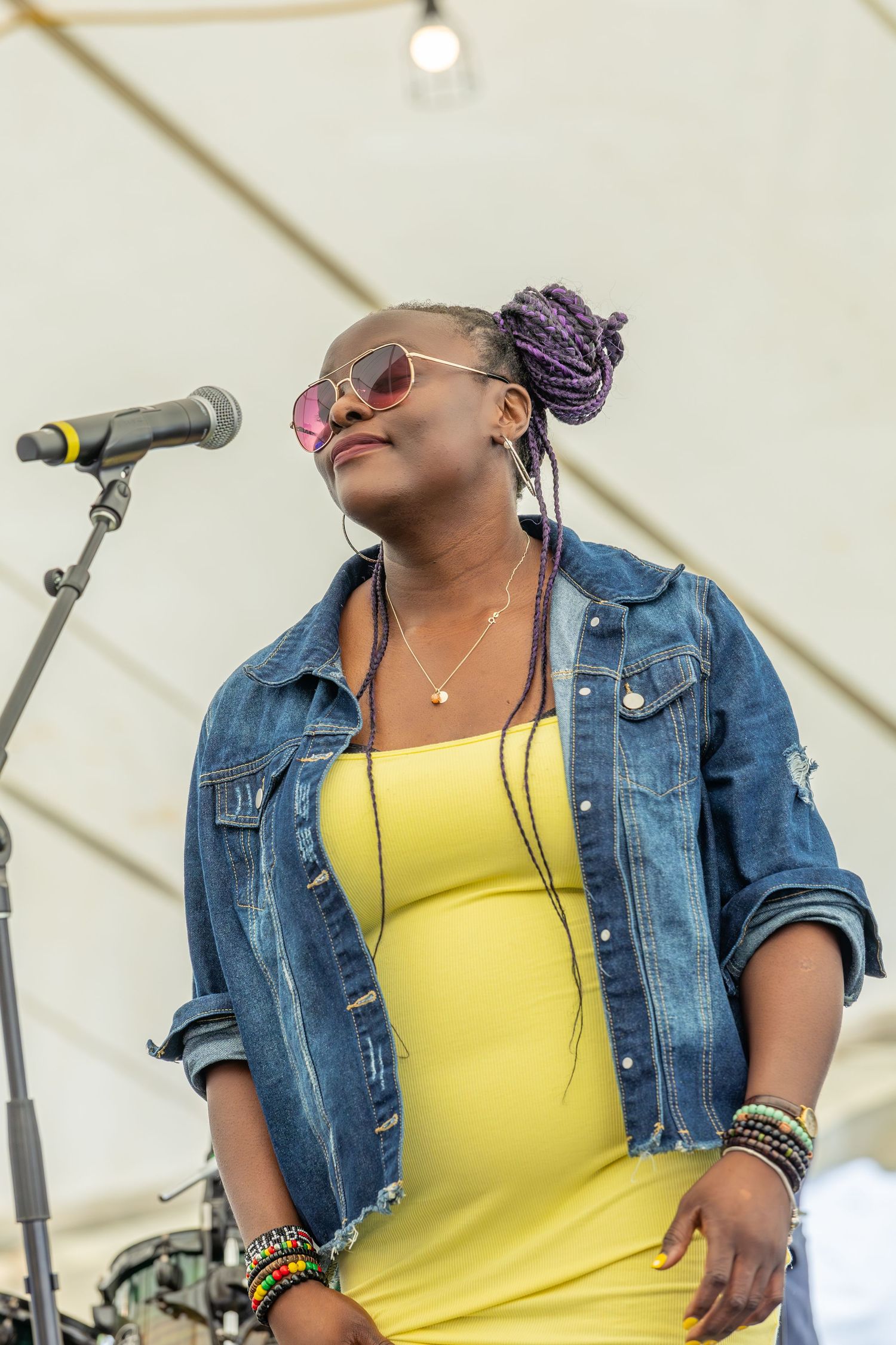 Singer in denim jacket and yellow top performing on stage with microphone during outdoor festival.