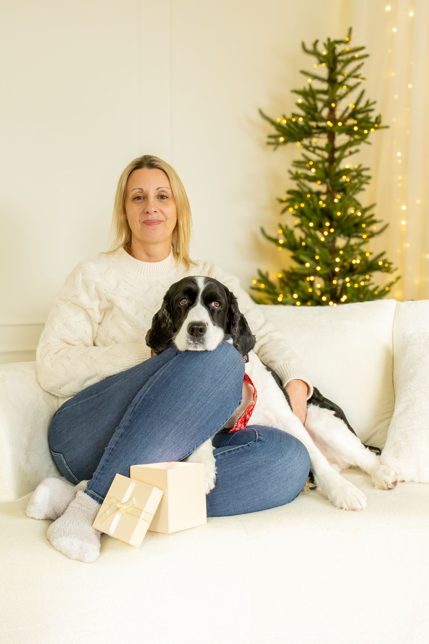 Smiling woman in a cream knit sweater sits on a white sofa with a black and white spaniel beside a lit Christmas tree and gift boxes.
