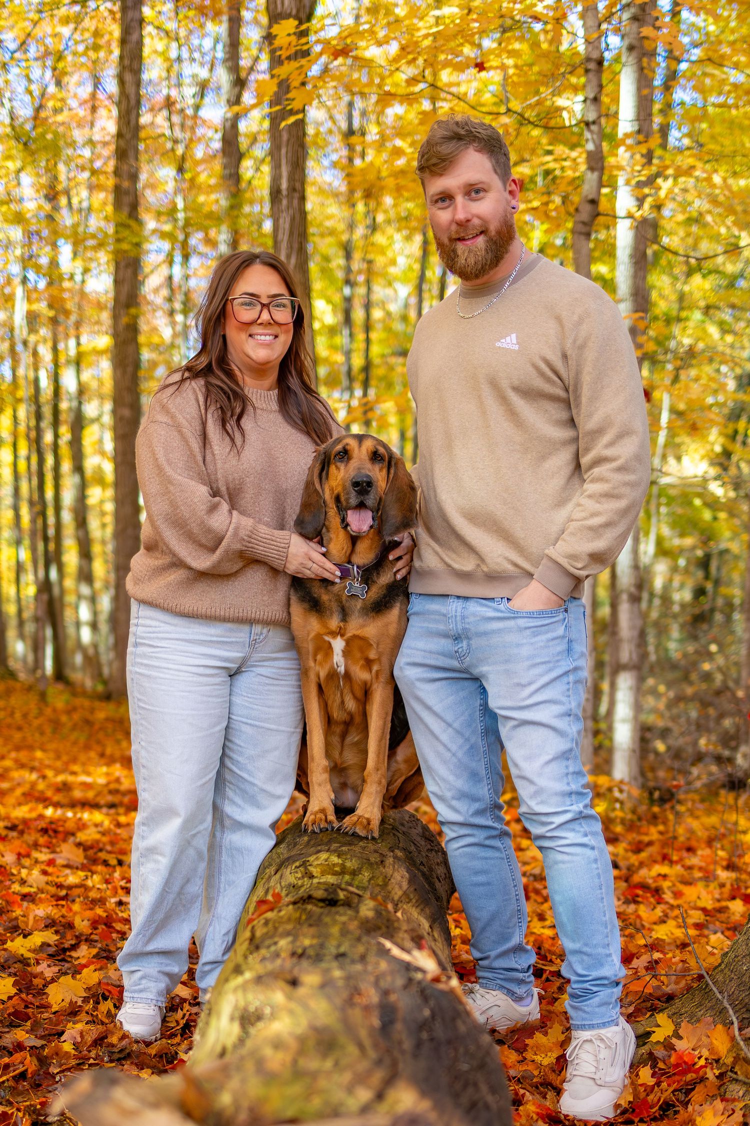 A couple in matching beige sweaters pose with their brown dog among vibrant fall foliage in the woods.