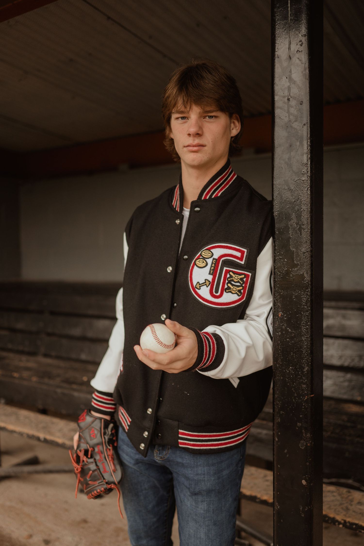 Baseball player poses with ball and glove wearing black and white varsity jacket.