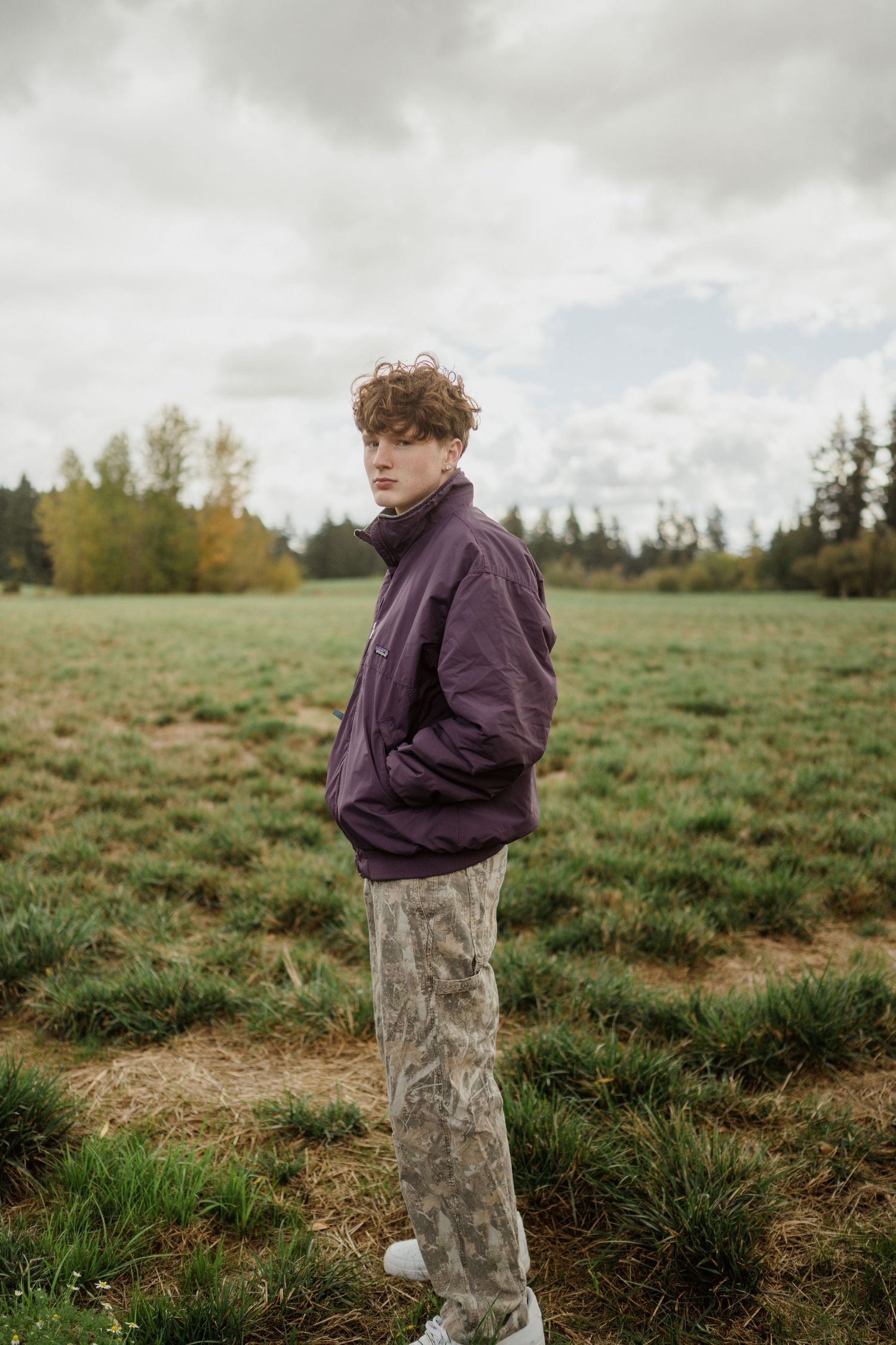 A person in a purple jacket and camo pants standing in a grassy field under cloudy skies.