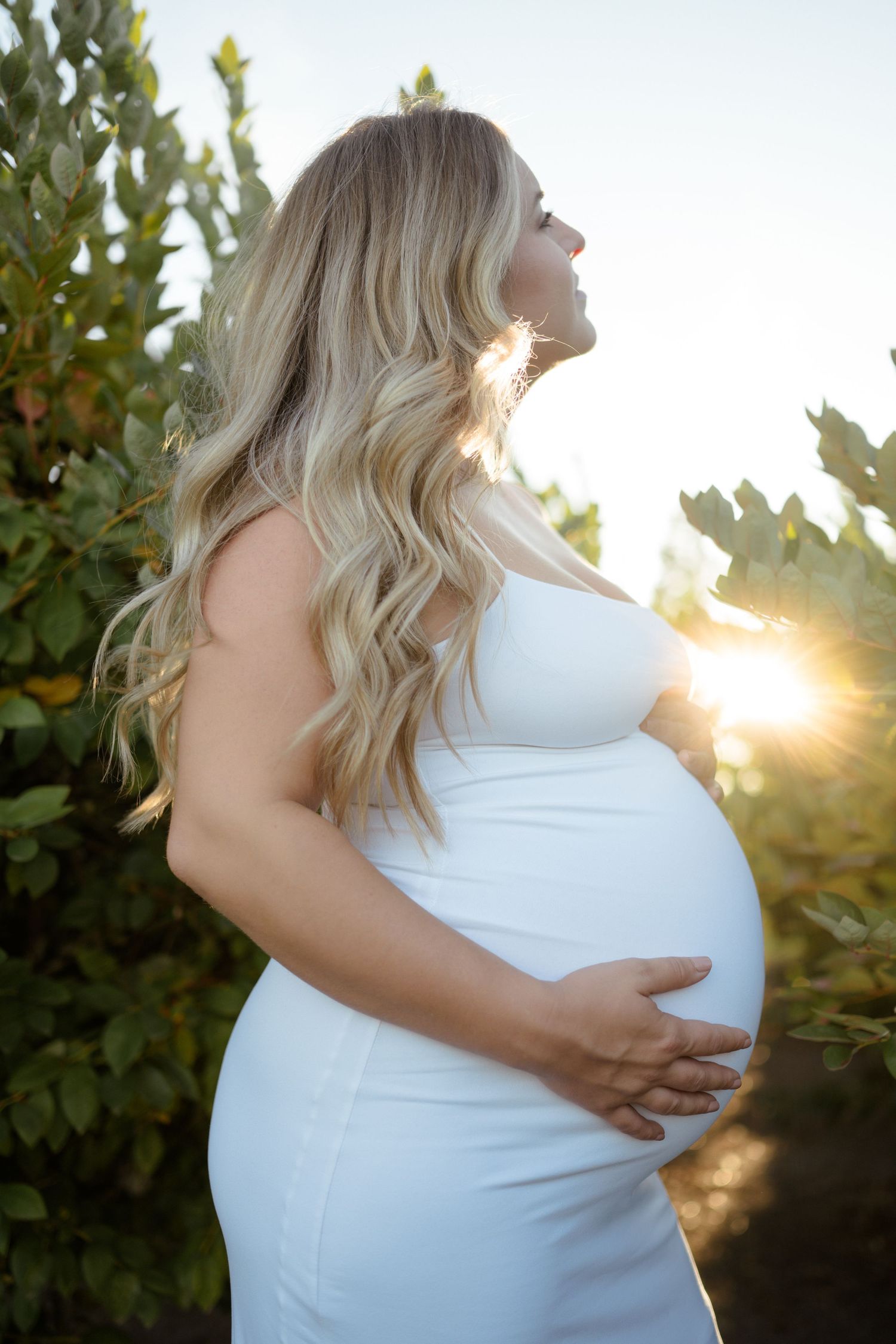 A pregnant woman in a white dress poses against sunlit foliage during a golden hour maternity photoshoot.
