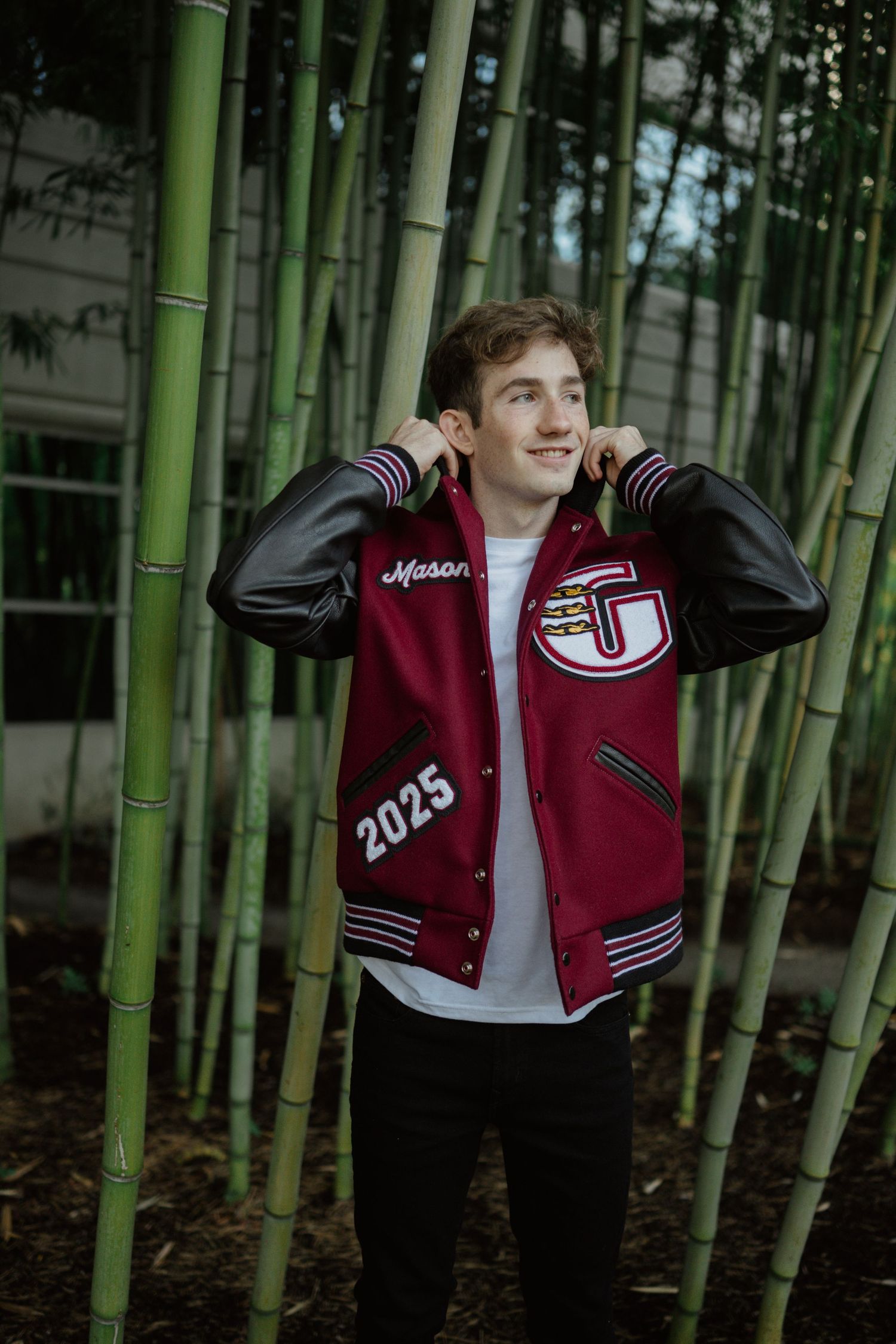 Student wearing varsity jacket poses in bamboo forest.