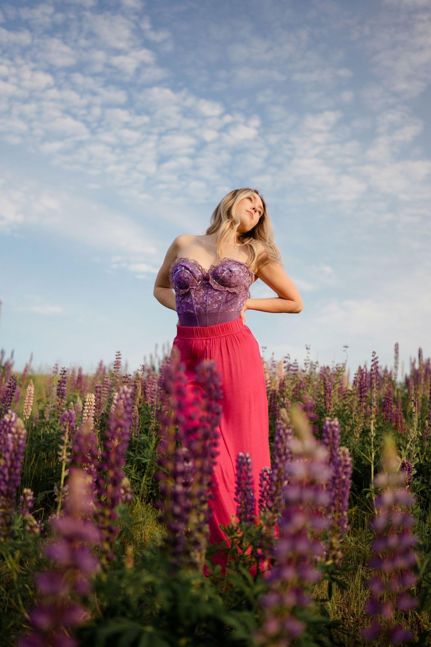 Woman in purple top and red skirt poses in field of blooming lupine flowers.