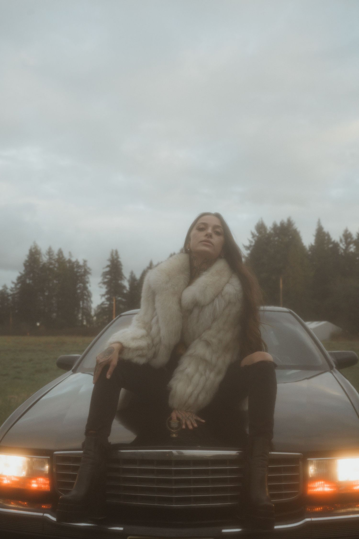 A person in a fur coat sits on the hood of a black car with headlights on at dusk with pine trees in the background.