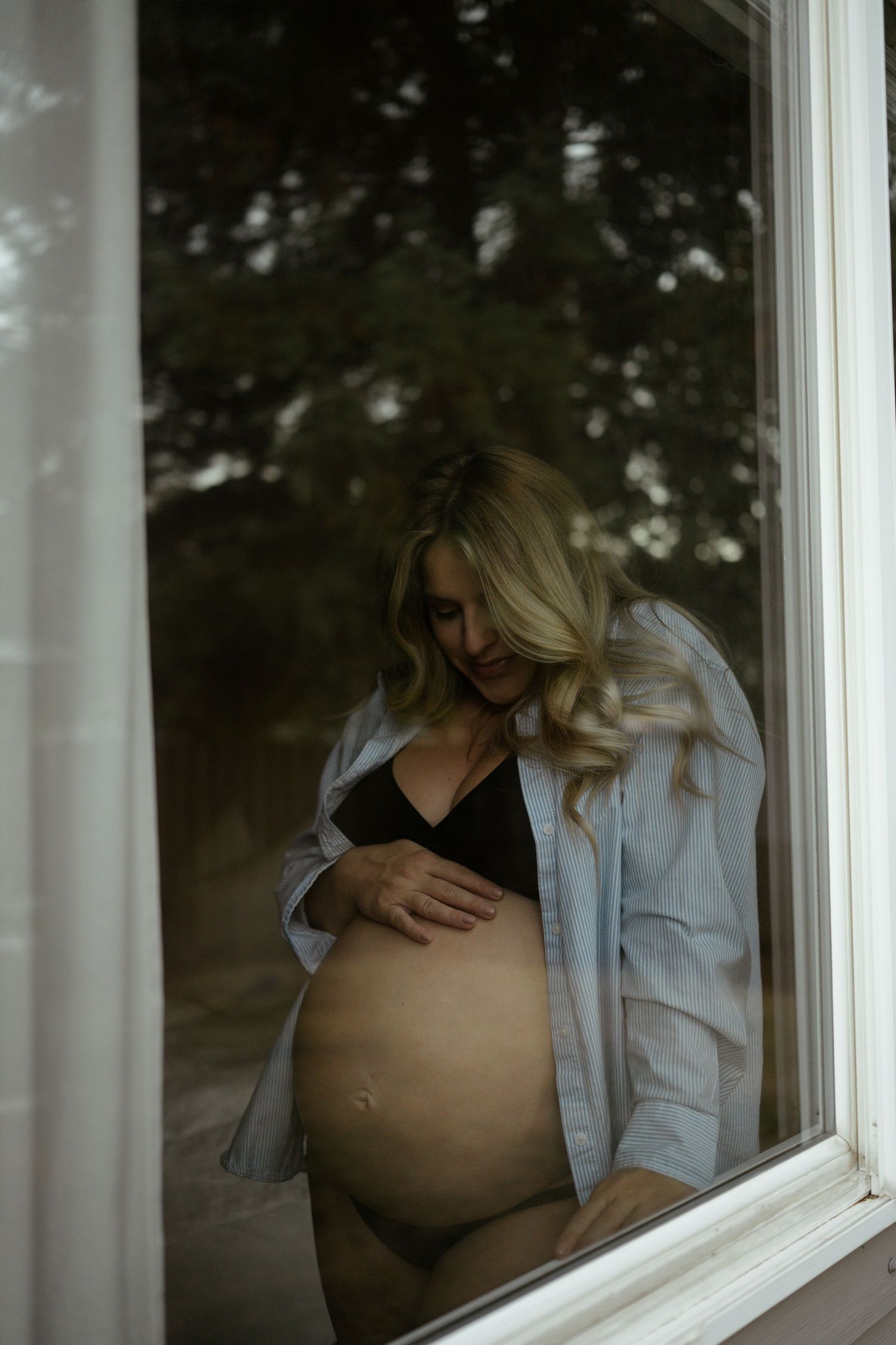 A maternity photo through a window shows an expectant mother in a denim shirt gently cradling her pregnant belly.