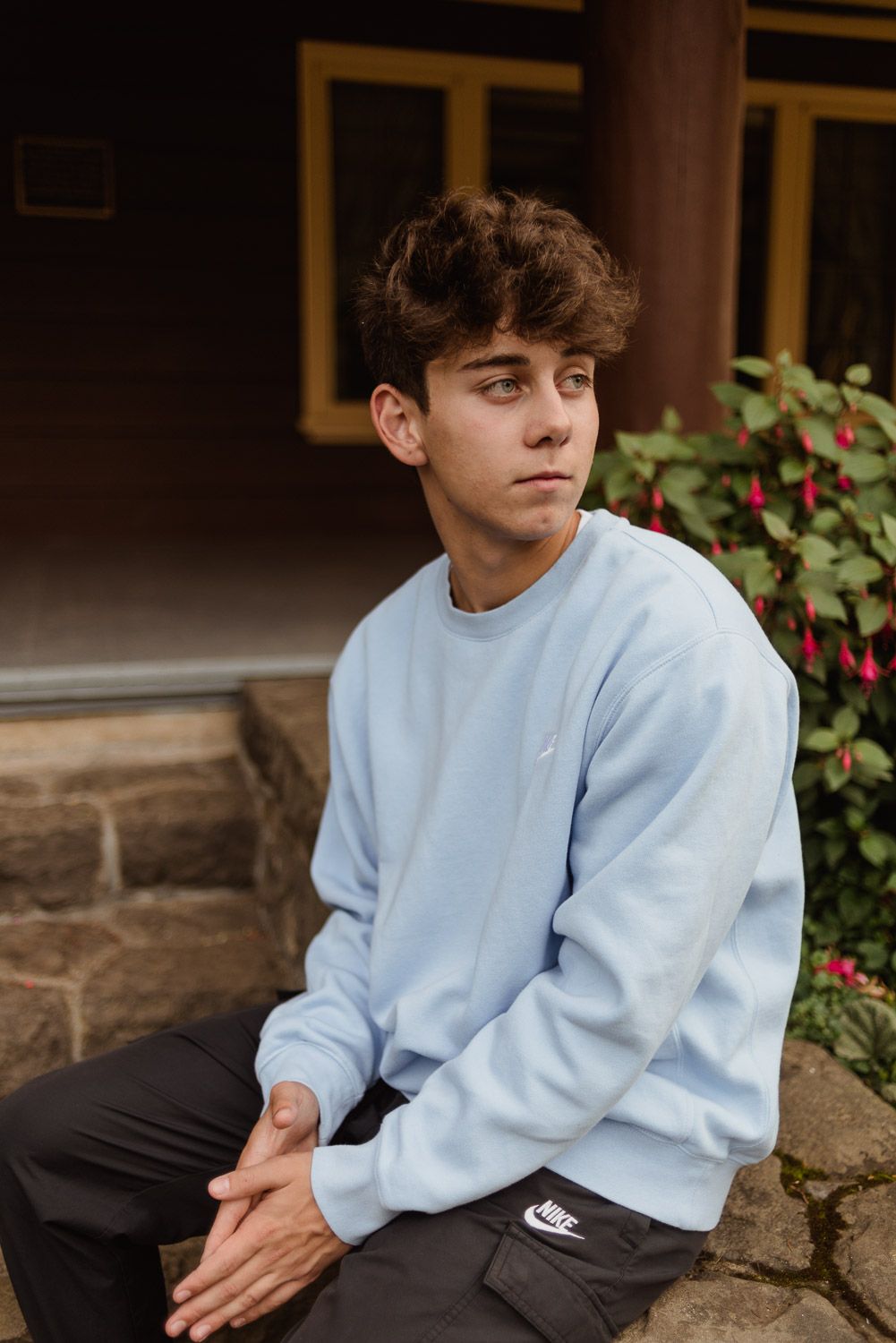 Young person in light blue sweatshirt sitting outdoors near flowering bushes.