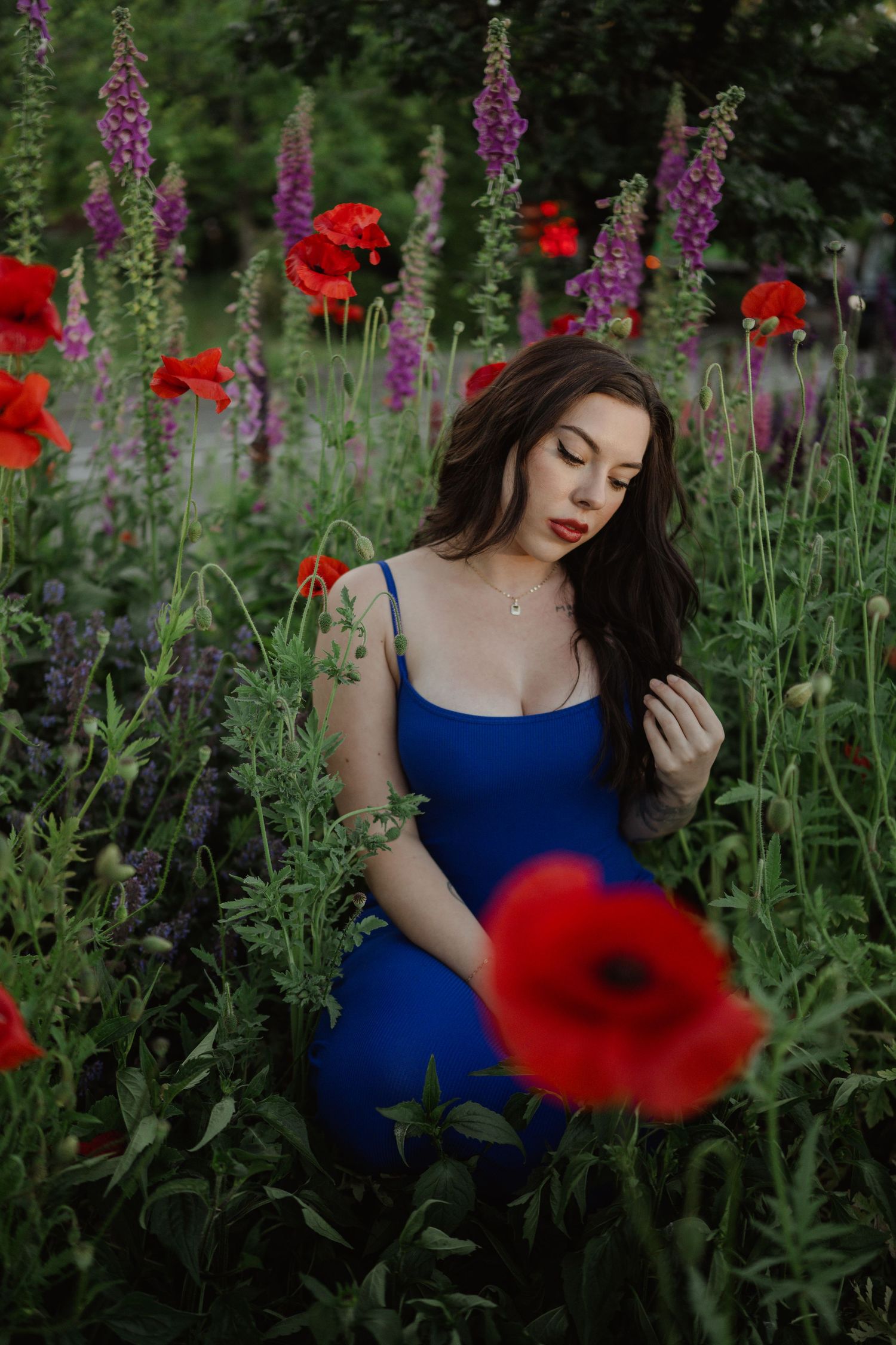 A person in a blue dress stands among vibrant red poppies and purple foxgloves in a lush garden setting.