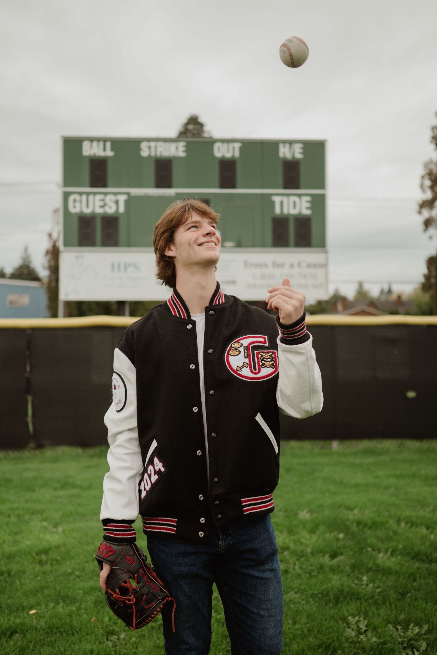 Baseball player tosses ball in air on field with scoreboard in background.