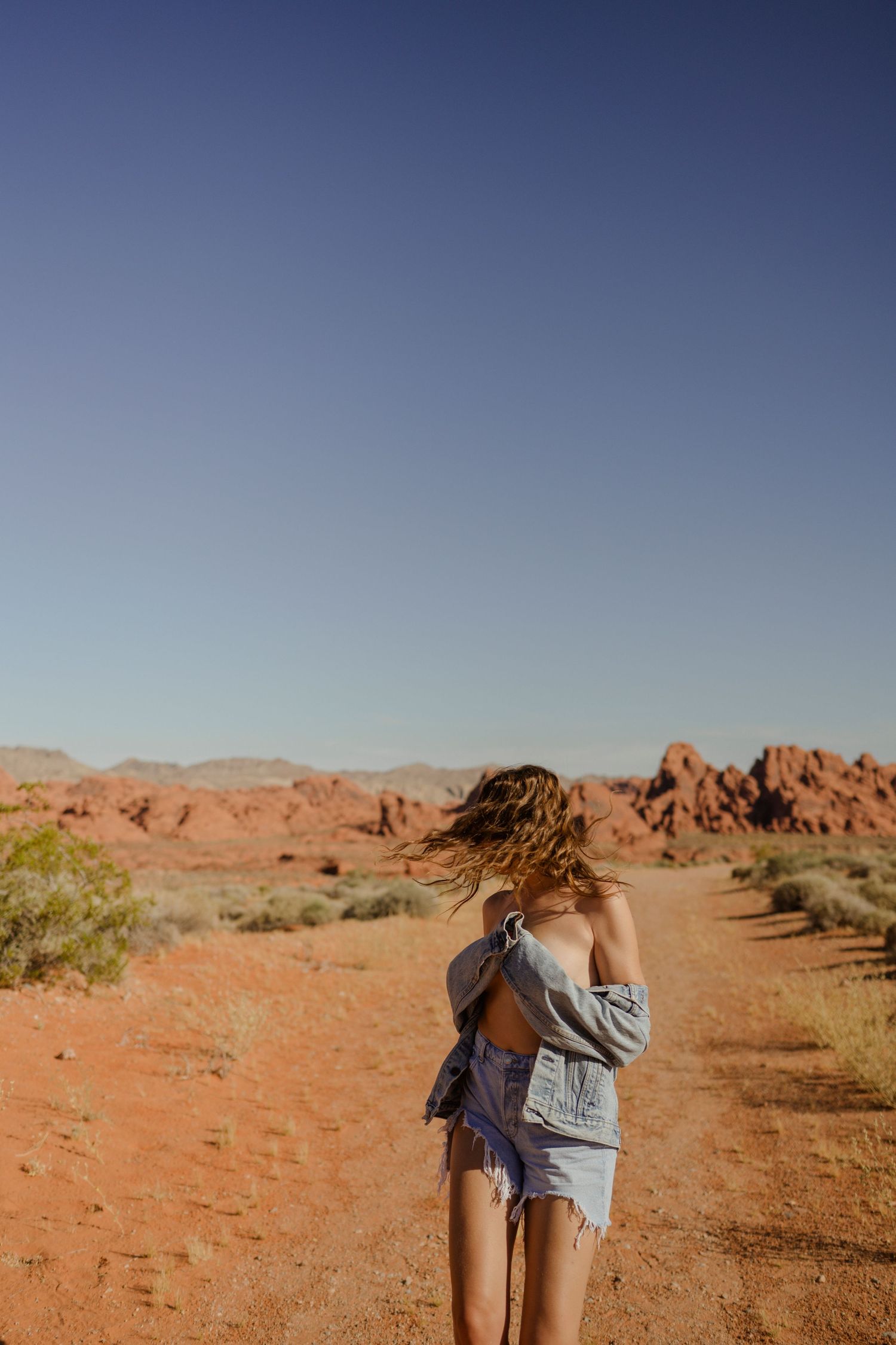 Person with windswept hair walking on red desert trail surrounded by rocky formations.