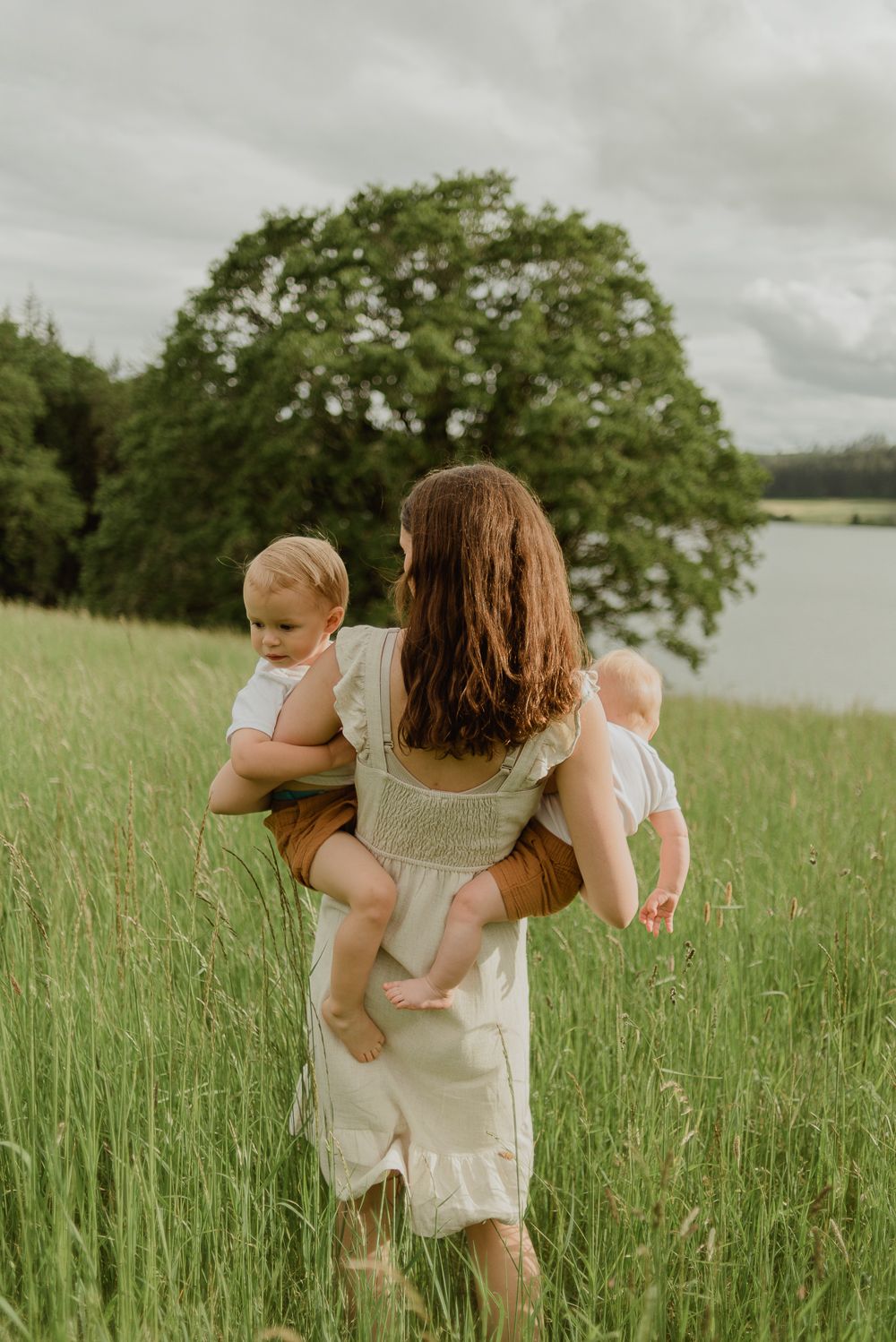 Woman in white dress carries children through tall grass by lake on cloudy day.