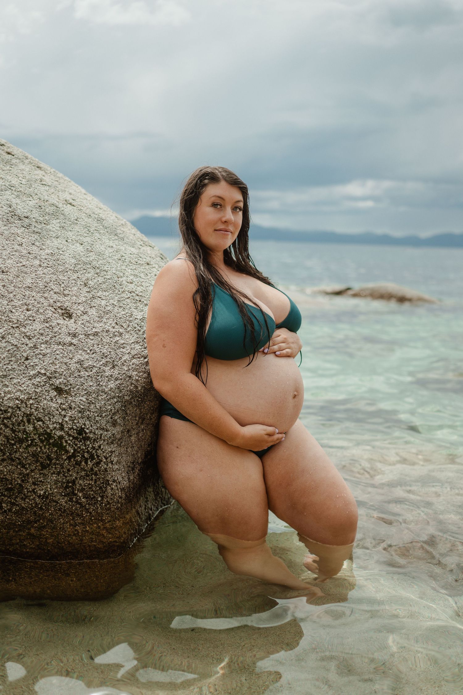 A person wearing a teal bikini poses in shallow tropical waters against a large granite boulder.