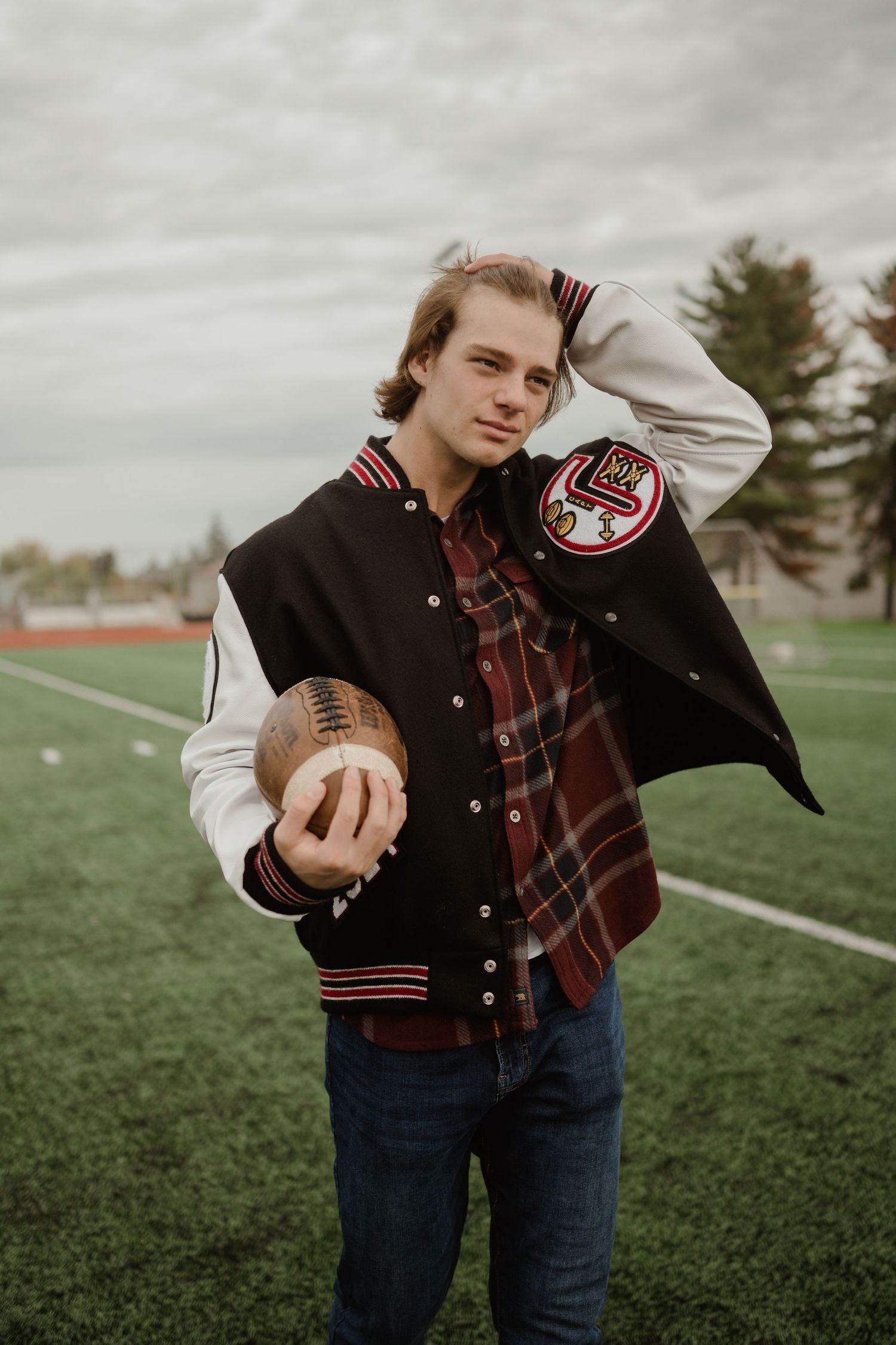 Baseball player holds football while wearing varsity jacket and plaid shirt on sports field.
