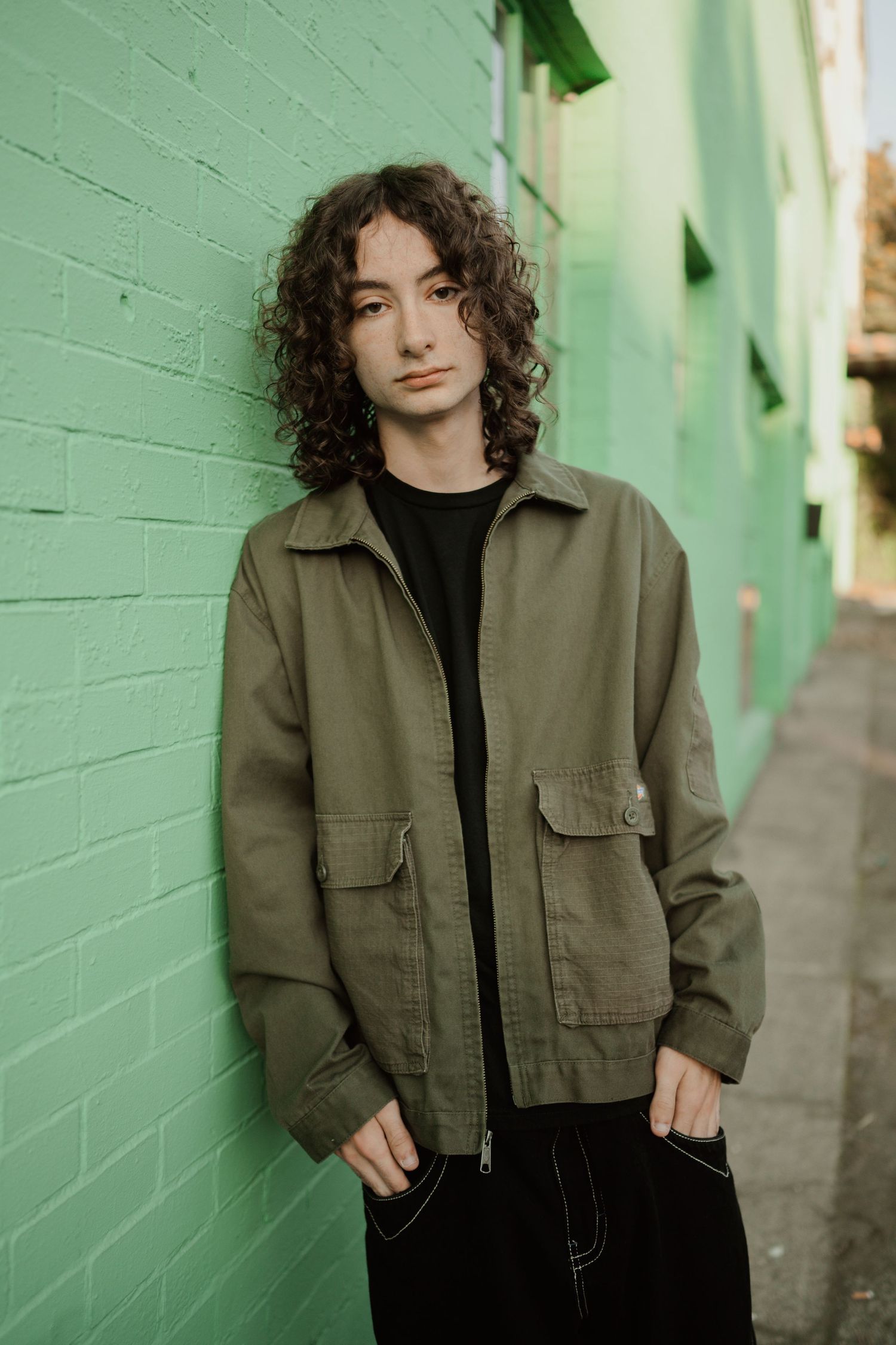 A portrait against a mint green wall wearing an olive utility jacket.