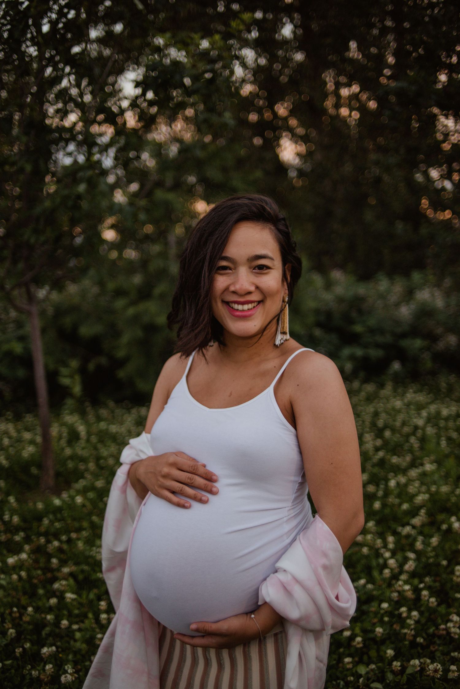 An expectant mother in a white tank top and light pink cardigan stands outdoors at sunset among greenery and trees.