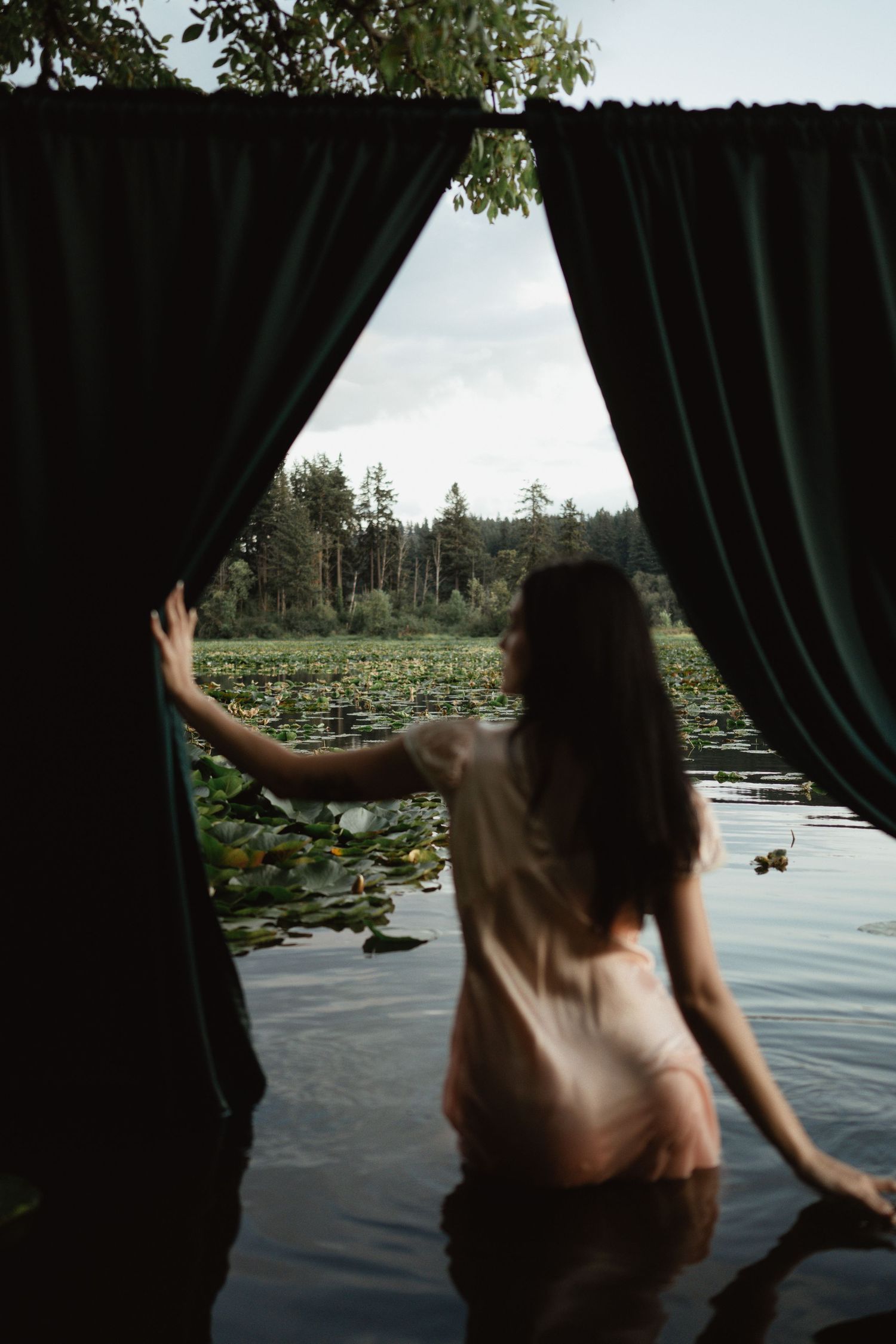 A figure in a sheer dress parts dark curtains to reveal a serene lake with lily pads and trees beyond.