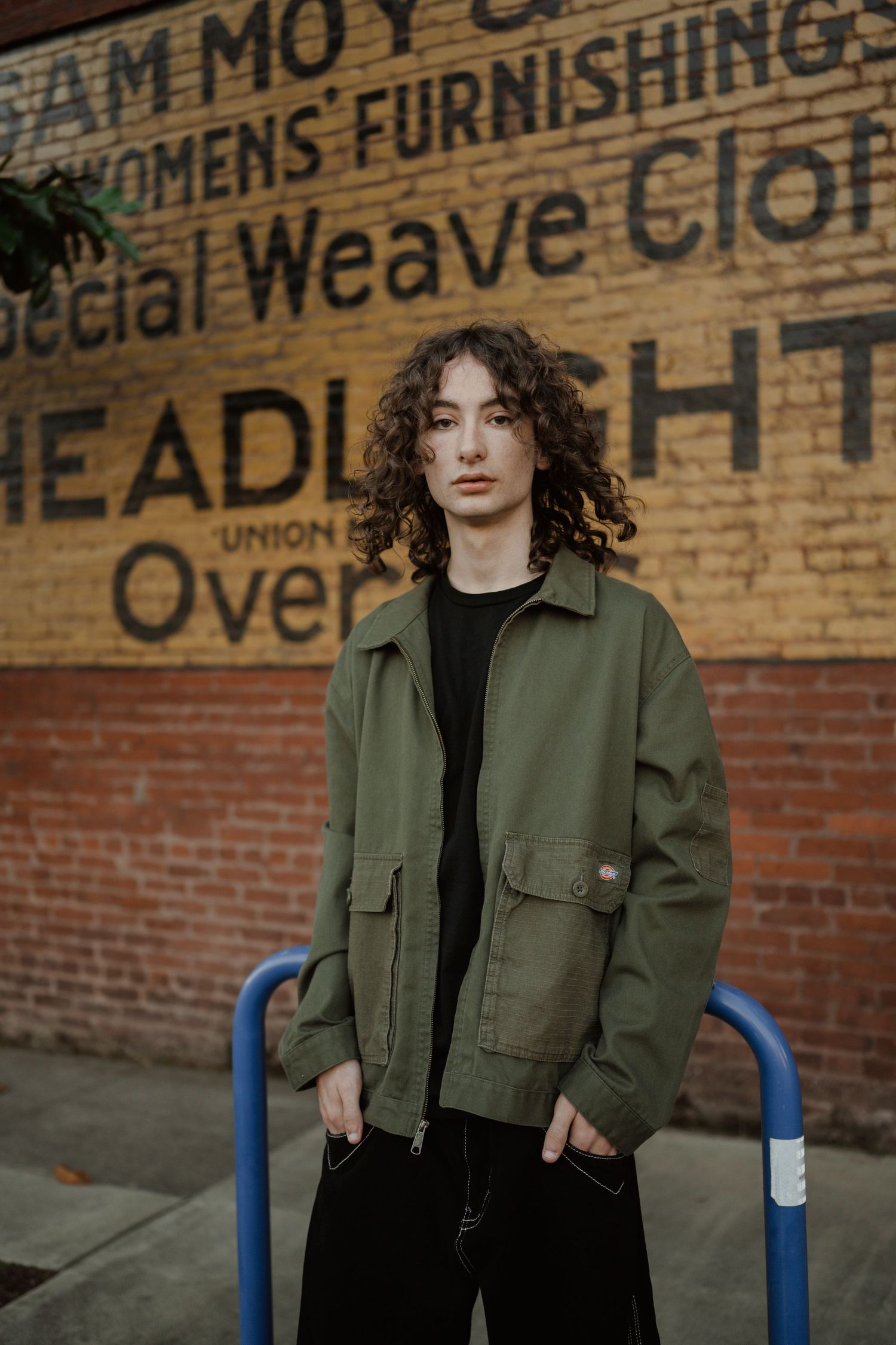A portrait against vintage wall signage wearing an olive jacket and black pants.