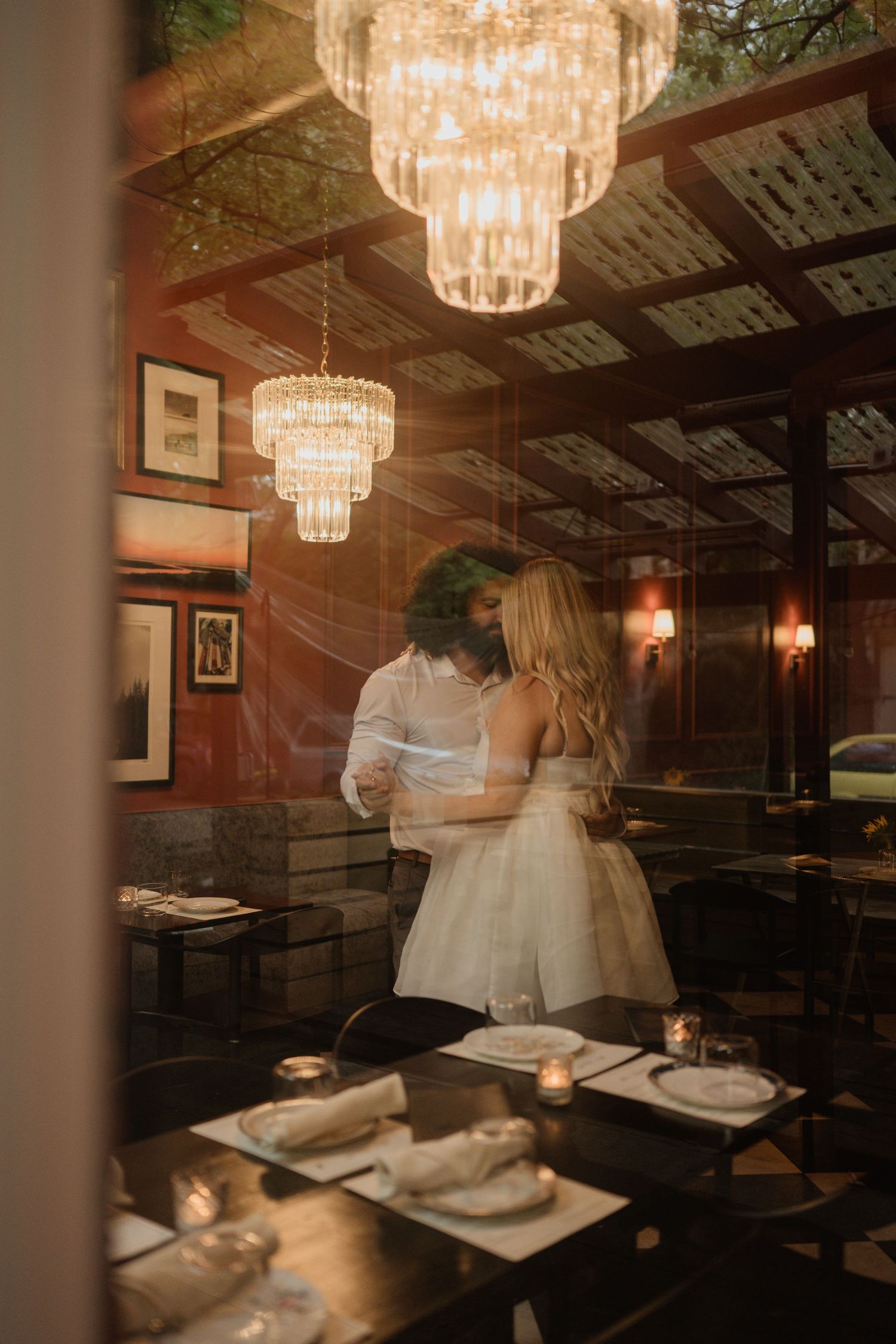 A couple embraces in a romantic restaurant setting beneath crystal chandeliers.