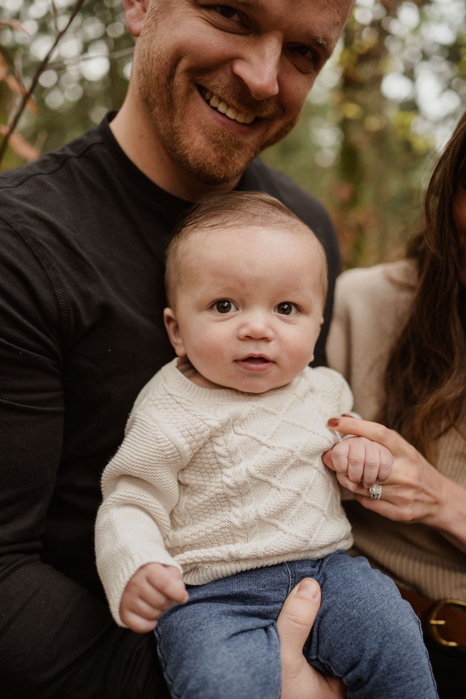 Close-up portrait of a baby in a white sweater with parents in a wooded setting.