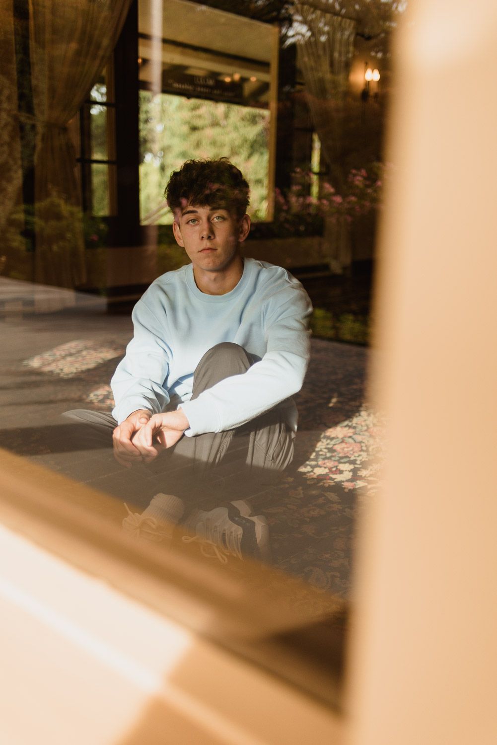Artistic portrait through window frame of person in light blue sweater sitting in serene setting.