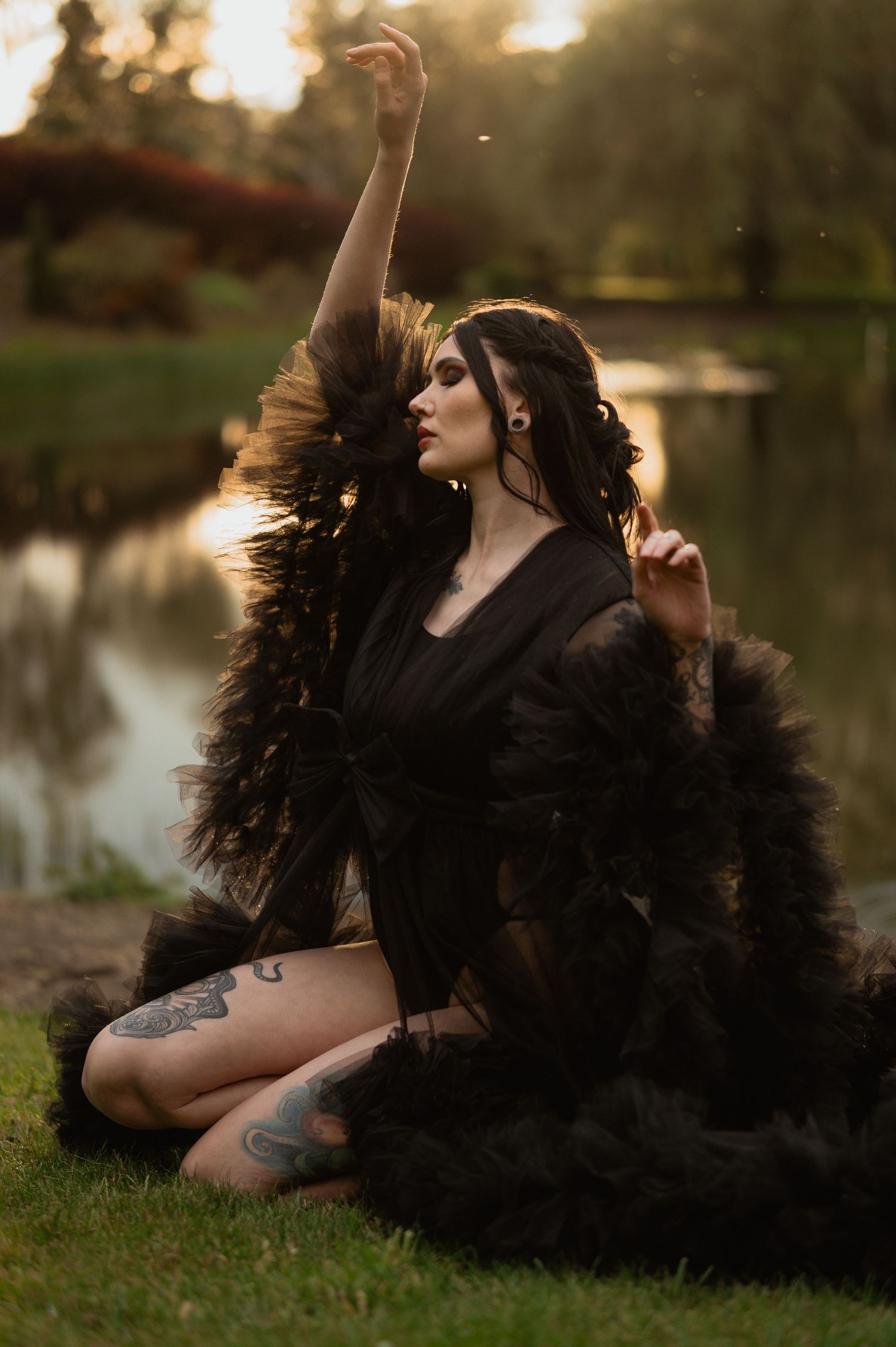 Moody artistic portrait of figure in black feathered dress posing gracefully by a pond at sunset.