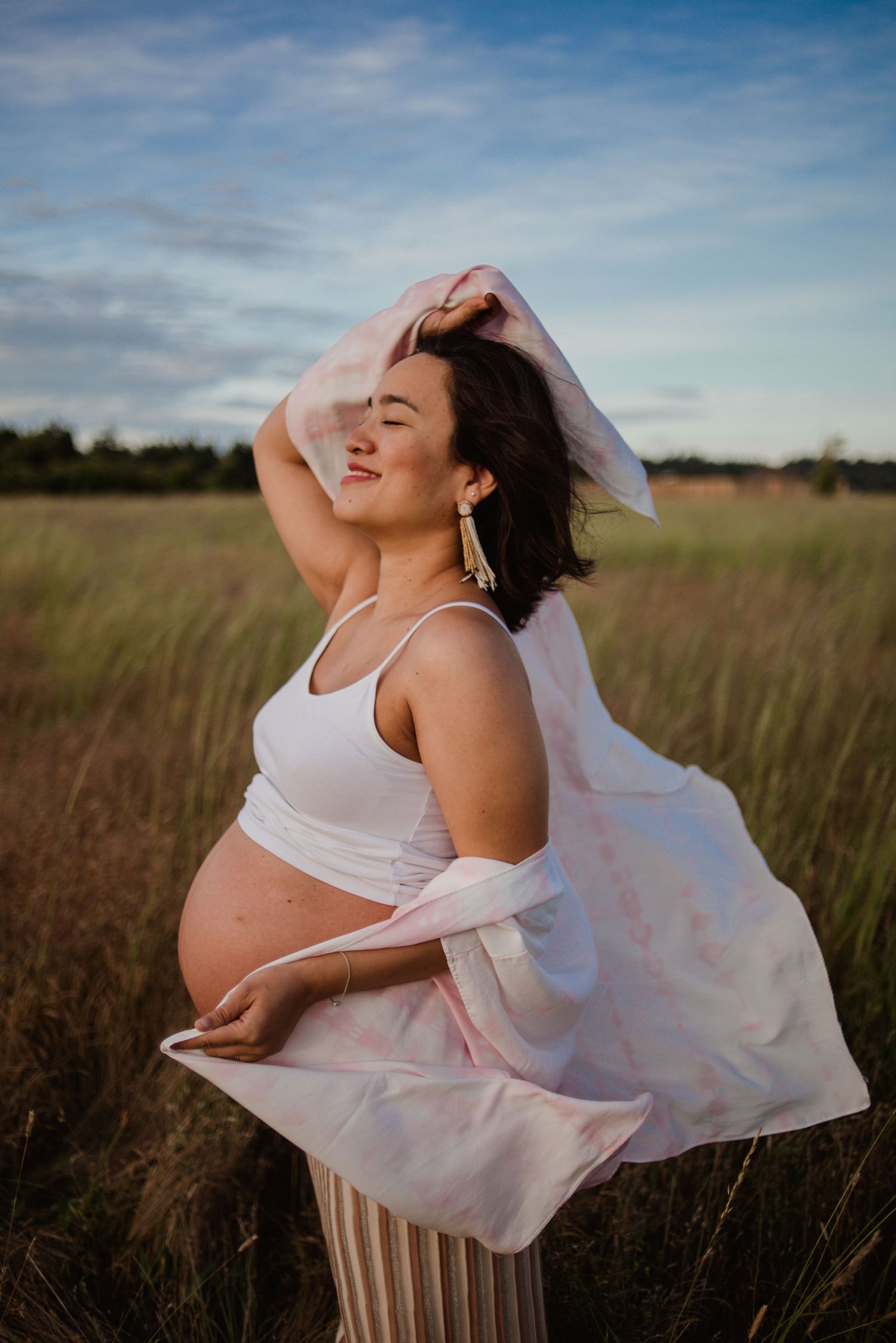 A pregnant woman in a white dress stands in a golden field at sunset with flowing fabric in the breeze.