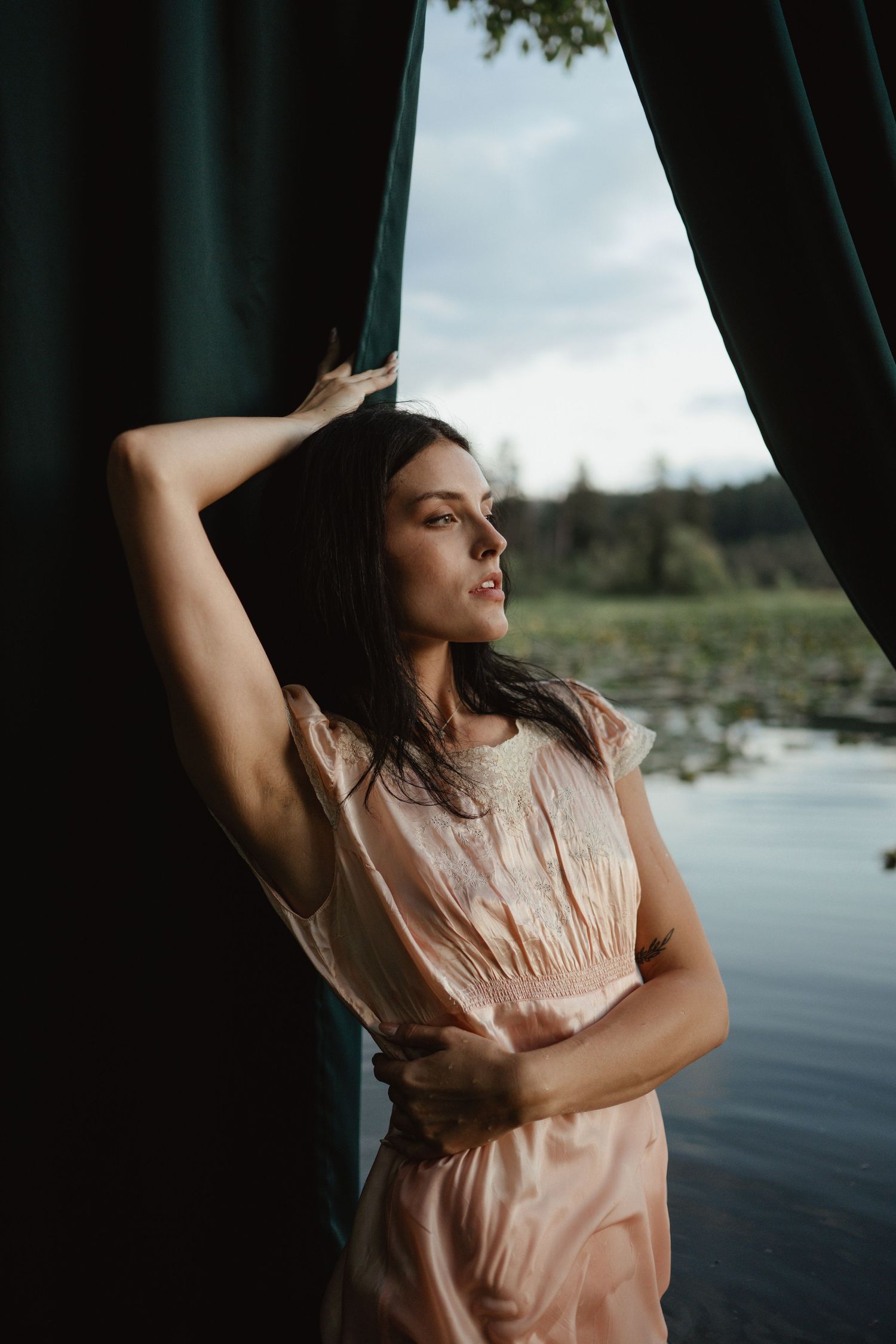 Figure in a peach-colored dress poses by a window against a scenic waterfront backdrop.