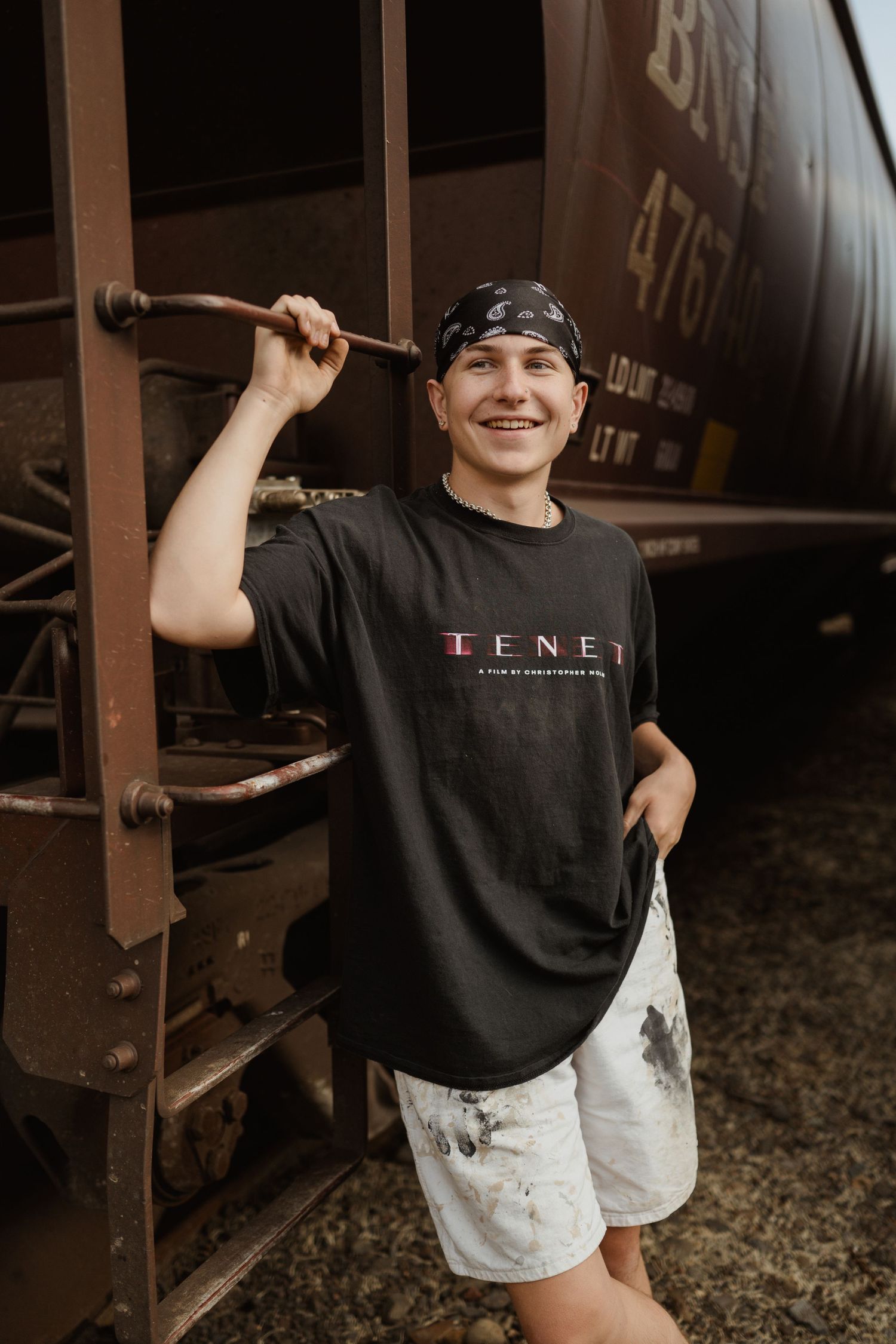 A casual portrait of someone in a TENET t-shirt and white shorts posing next to a rustic train car.