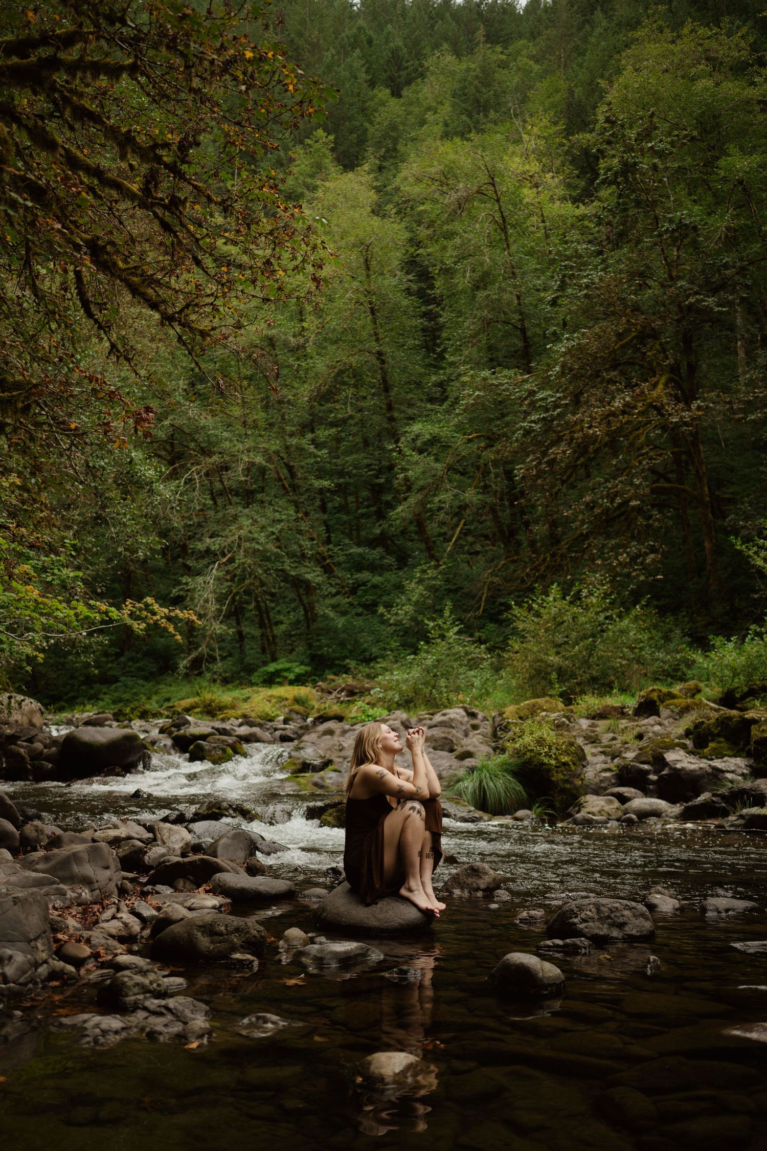 Person sitting on rocks by forest stream in moody natural setting.