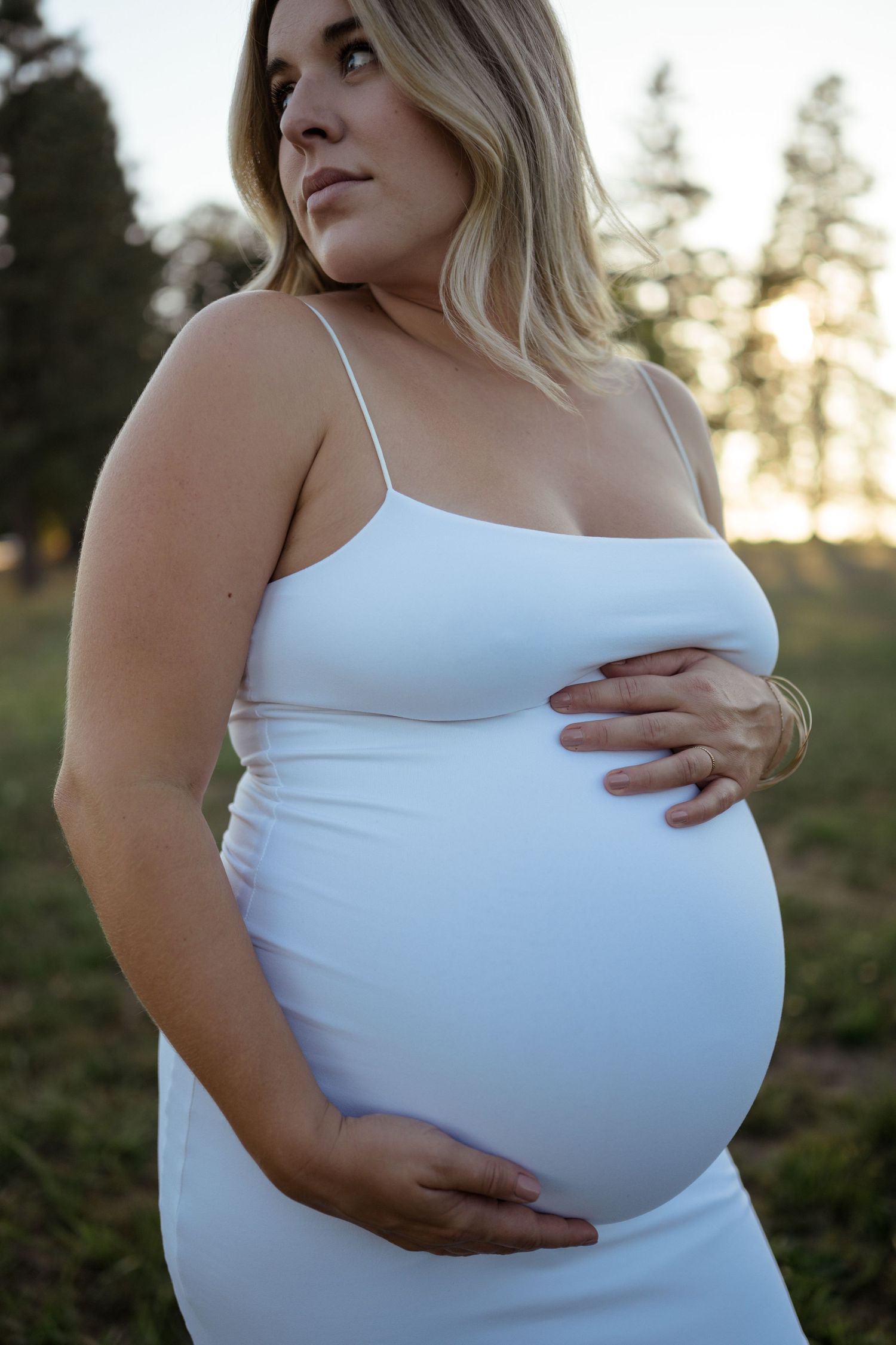 Expectant parent wearing white dress cradling baby bump during golden hour sunset outdoors.