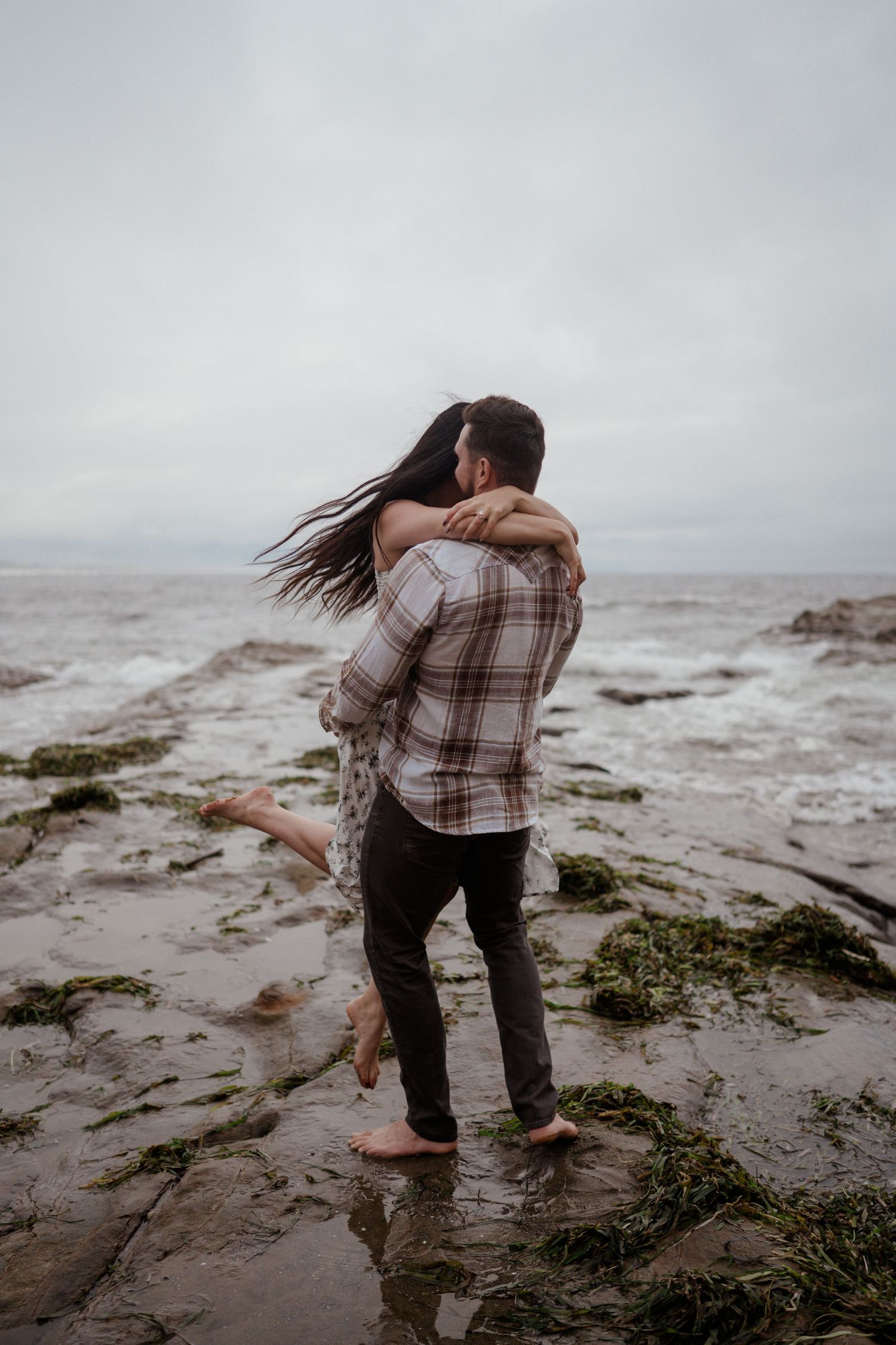A couple embracing on rocky coastal shores during an overcast day.