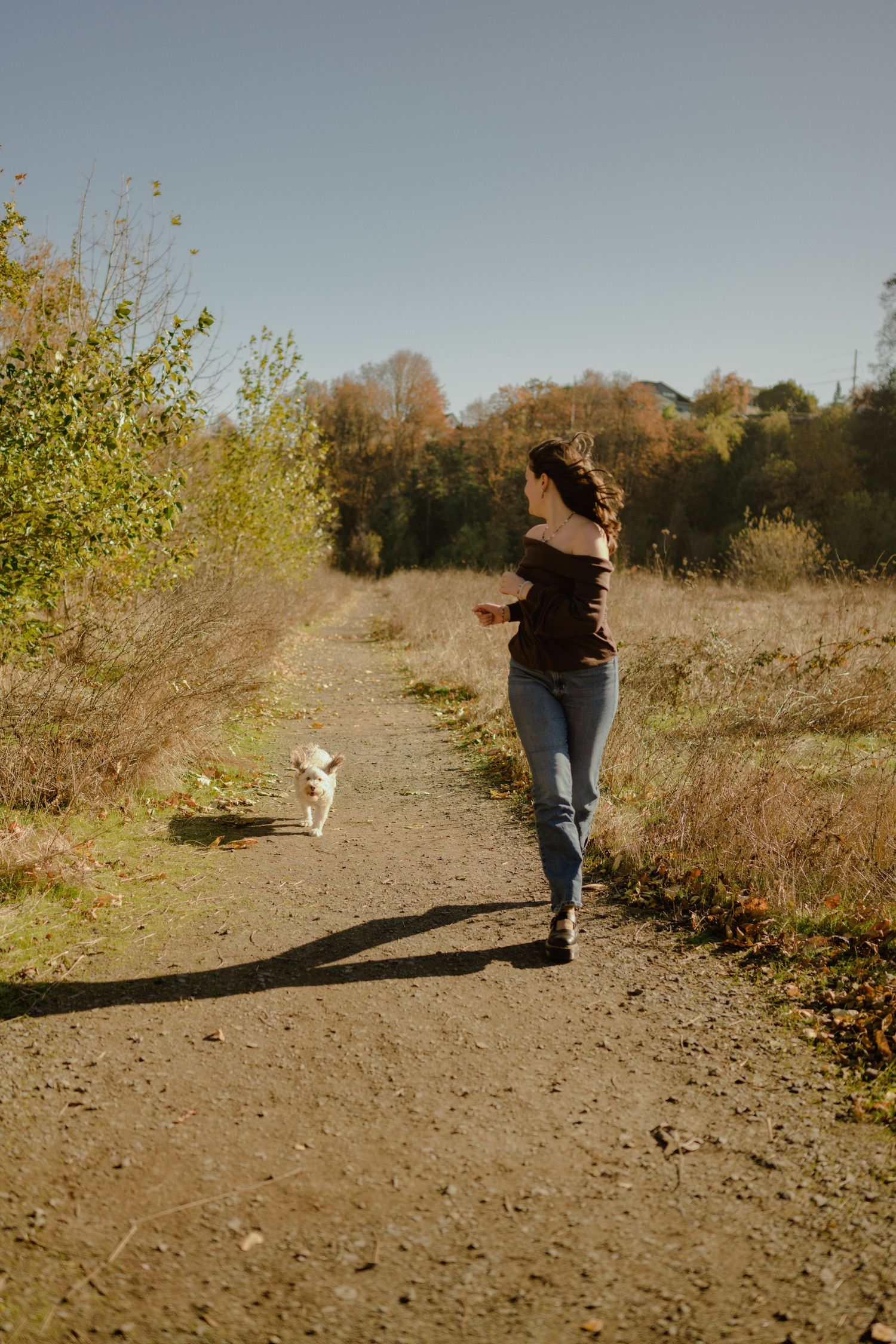 A person walks along a sunny autumn trail with a small white dog.
