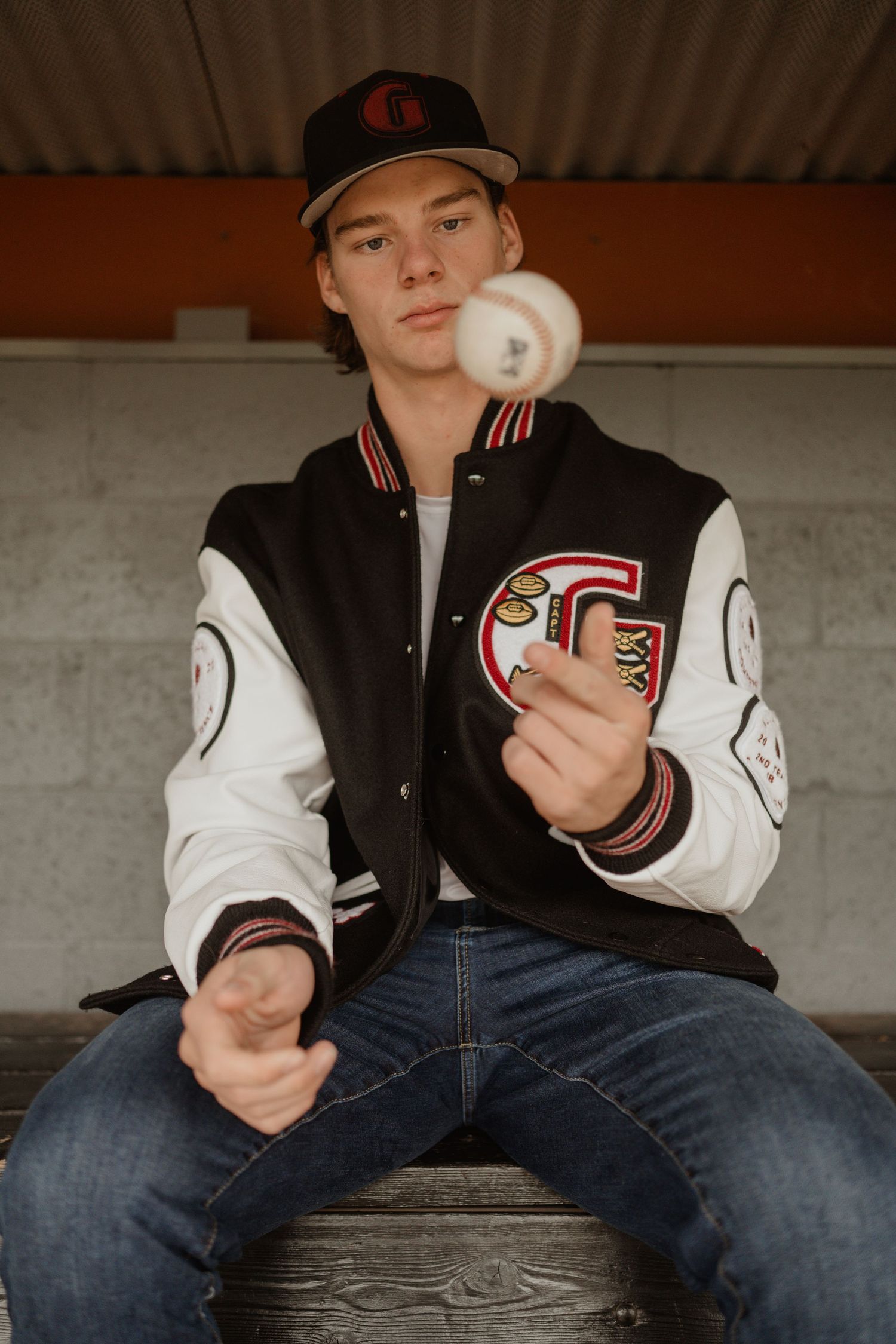 Baseball player in varsity jacket tosses a ball while sitting on wooden bleachers.
