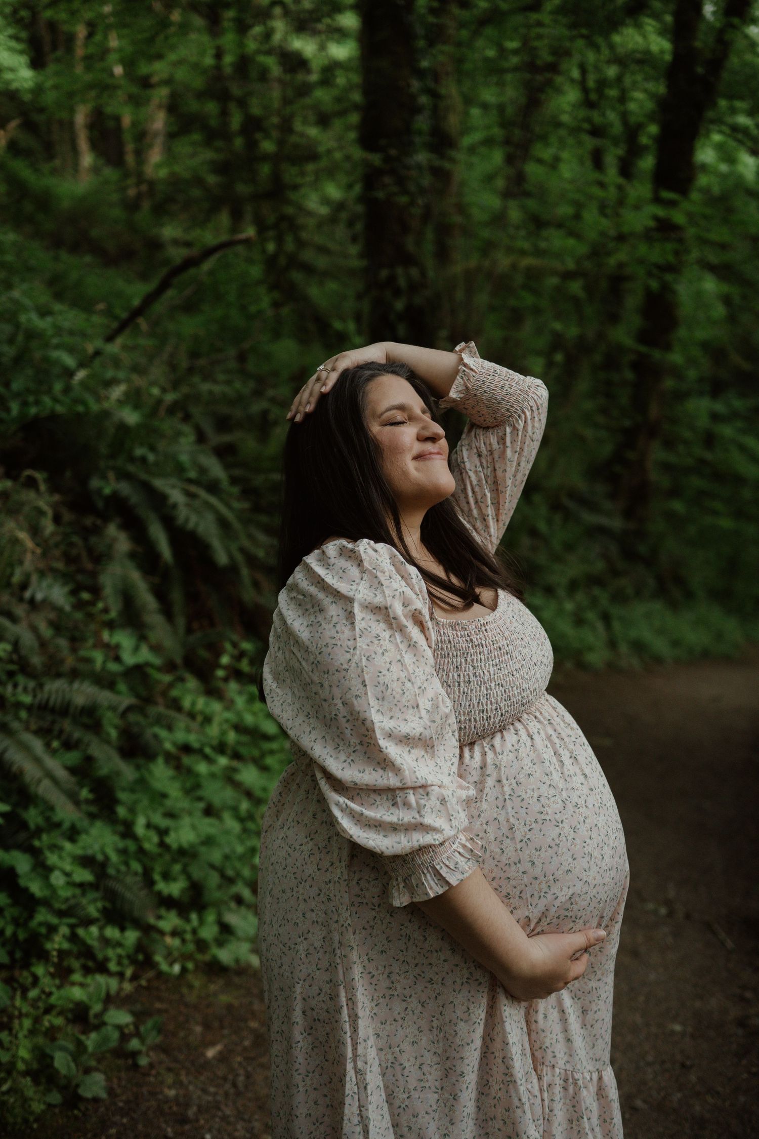 Pregnant woman in flowing cream dress poses serenely on forest path surrounded by lush green trees.