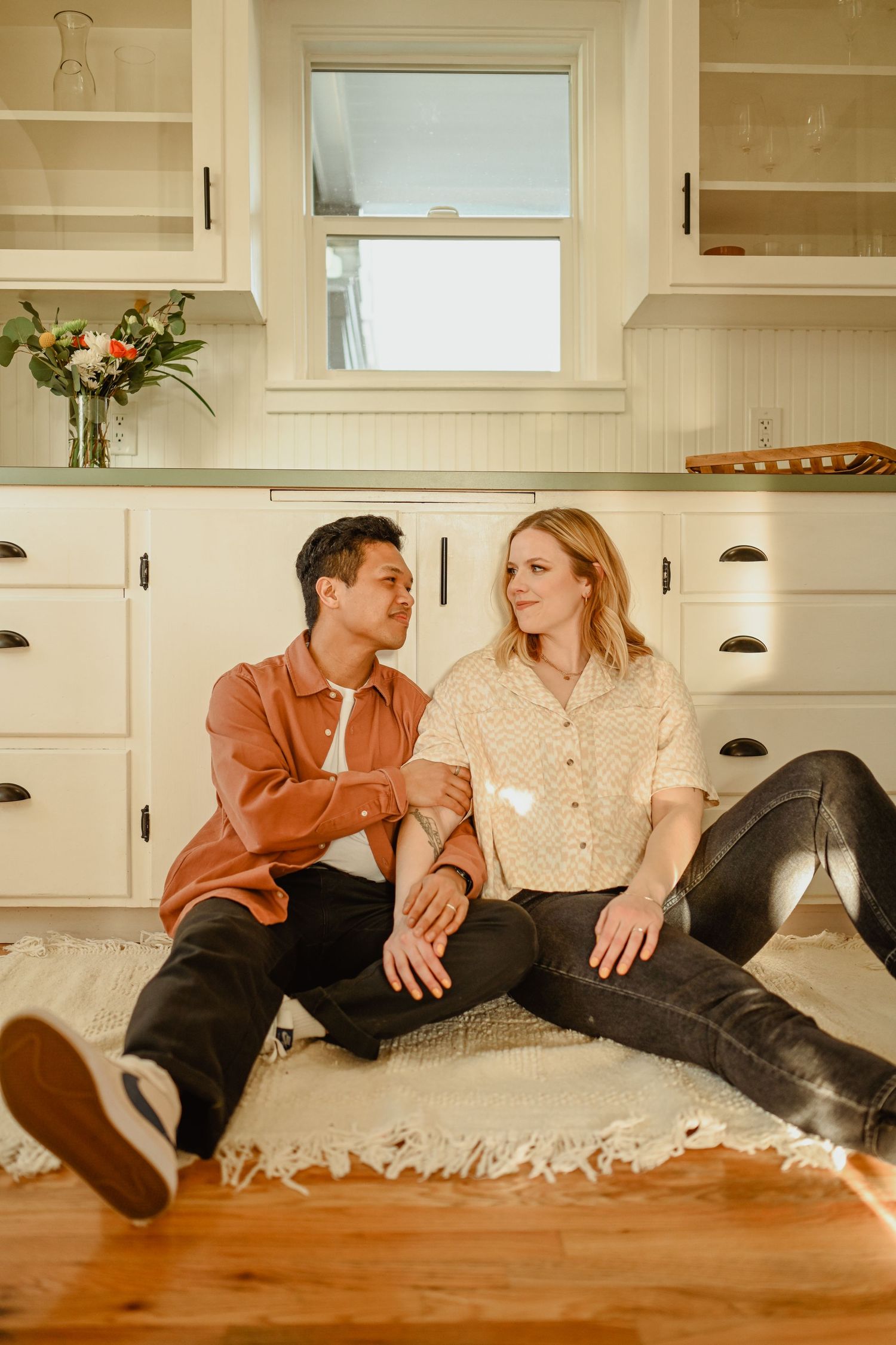 A couple sitting casually together on a kitchen floor with white cabinets and hardwood.