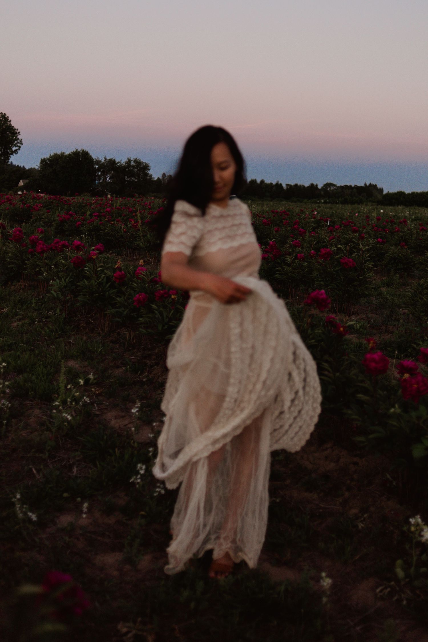 Figure in a flowing white lace dress stands in a garden of red roses at dusk with a pink-tinged sky.