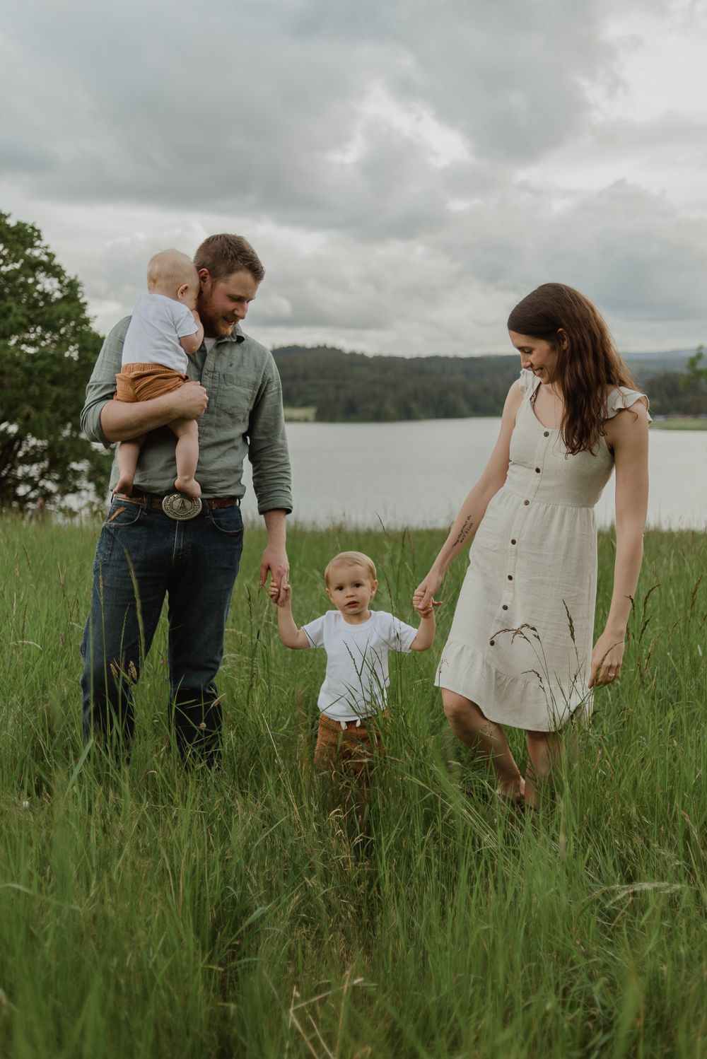 Family walking through tall grass meadow near lake on overcast day.