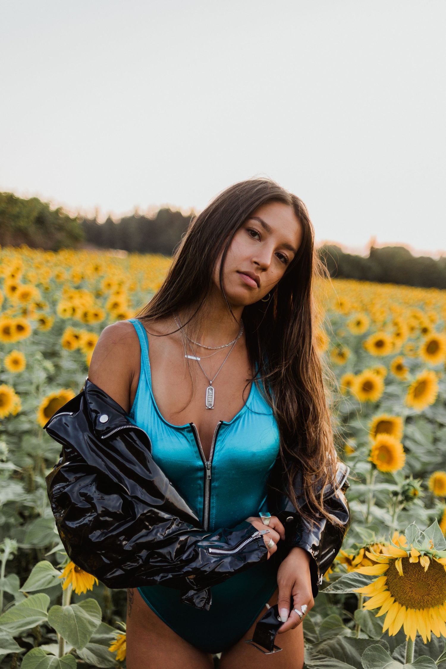 A person poses in a turquoise top and black jacket among vibrant yellow sunflowers during golden hour.