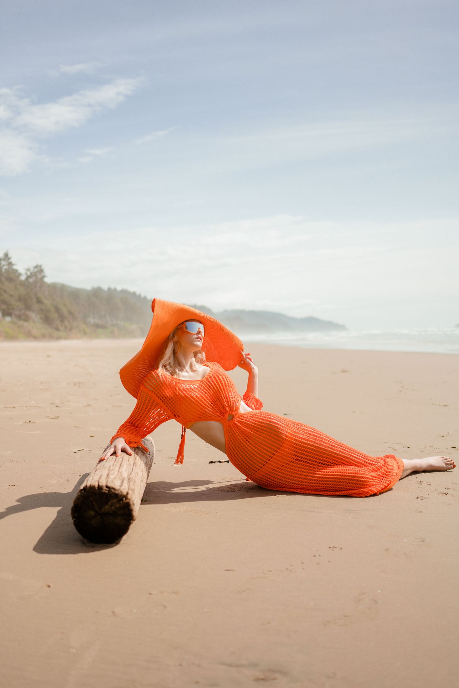 Person in bright orange sweater dress lying artistically on driftwood at beach.
