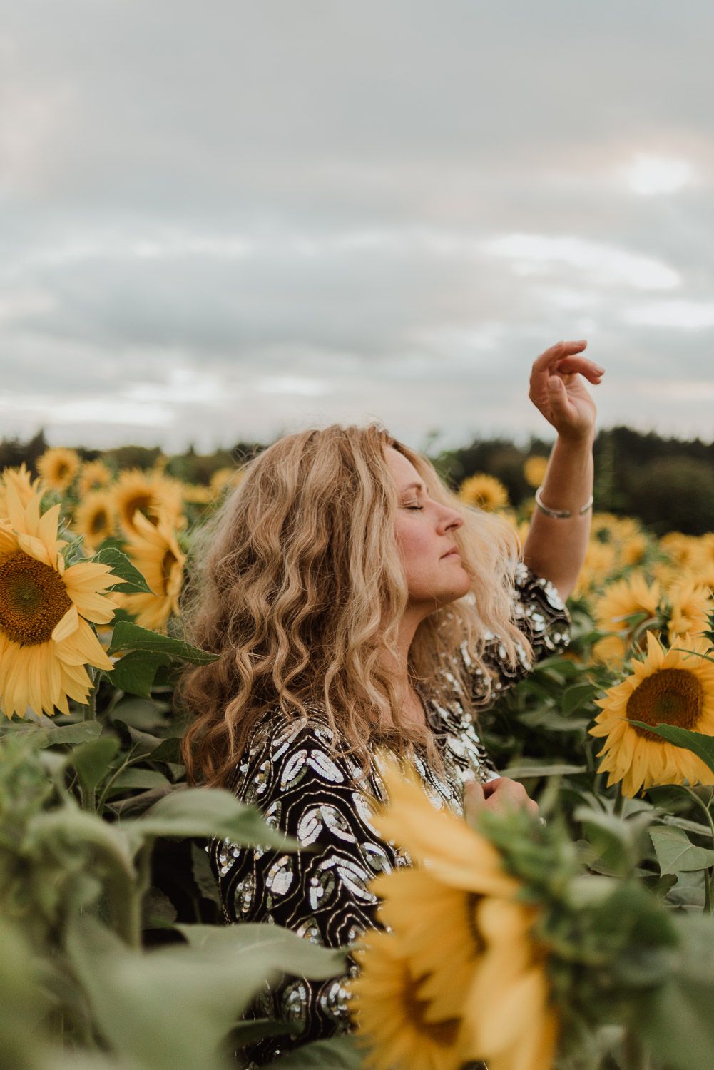Woman with curly hair poses dreamily in a vibrant sunflower field under cloudy skies.