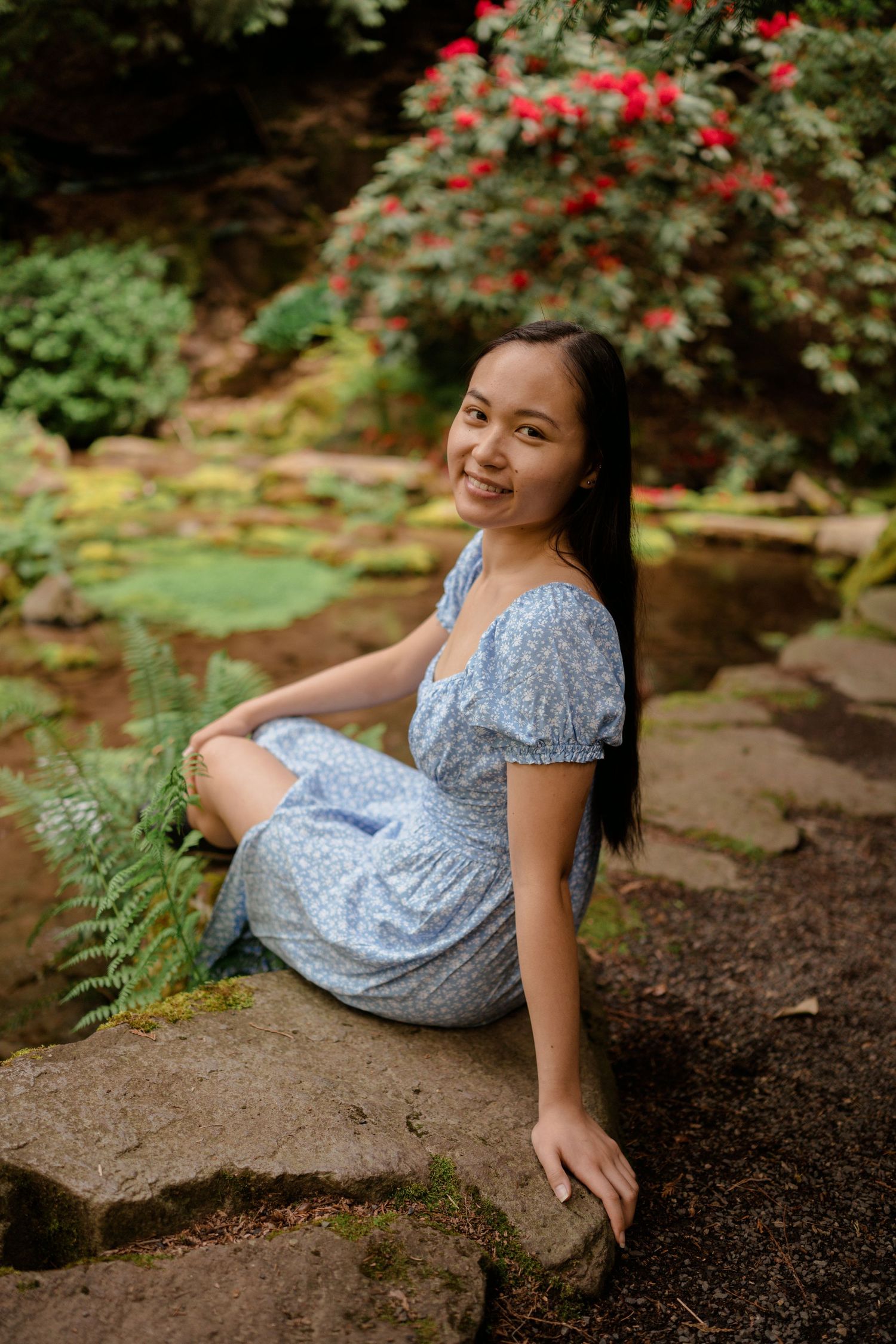 Garden portrait in blue floral dress sitting on stone surrounded by lush greenery.