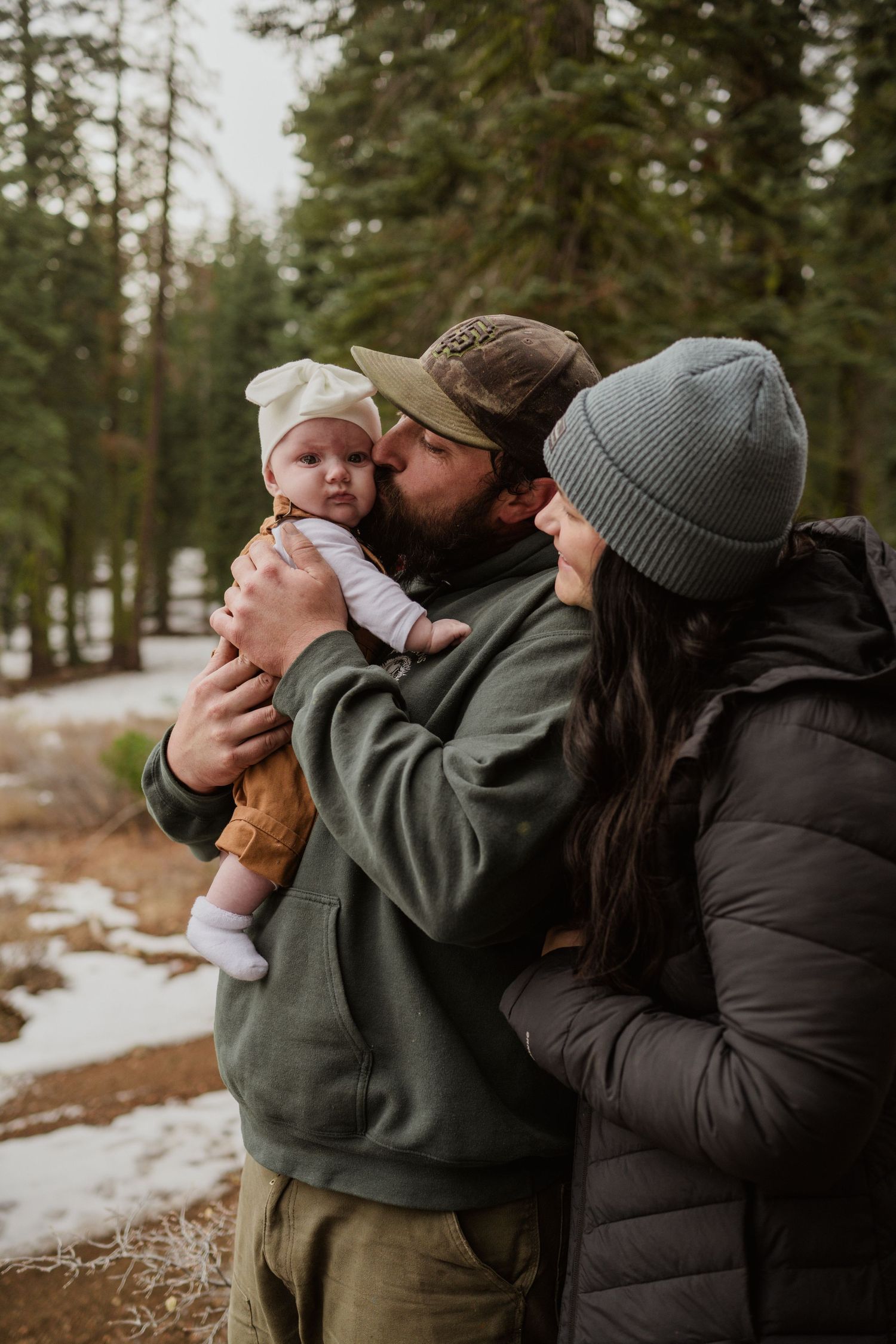 A tender family moment captured in a snowy forest setting with warm winter clothing and evergreen trees.