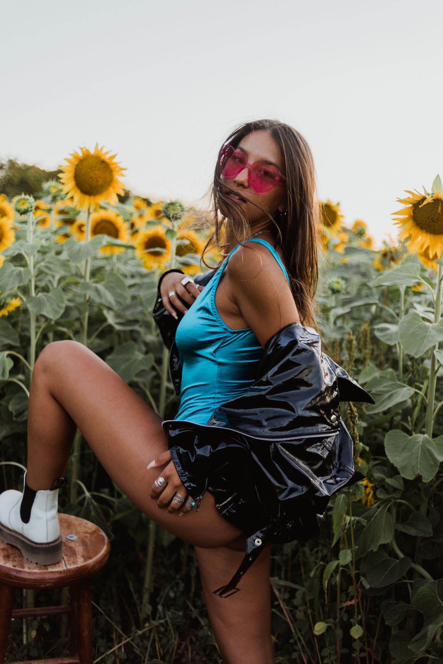 A person in pink sunglasses and turquoise tank top poses playfully in a vibrant sunflower field at sunset.