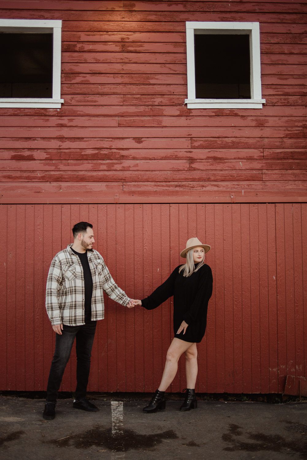 A stylish couple posing playfully against a red barn wall with white-trimmed windows.