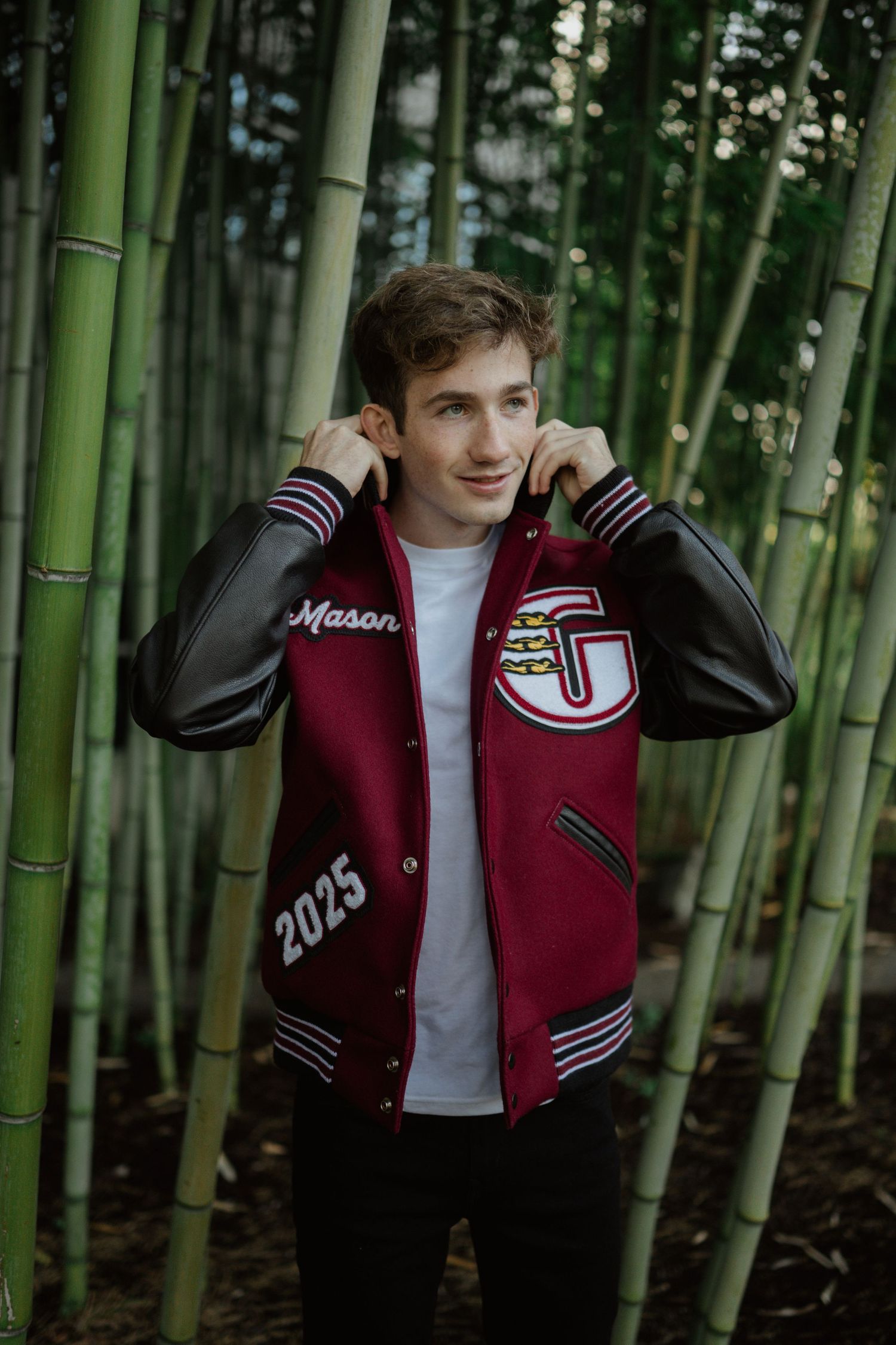 Person wearing maroon varsity jacket with '2025' lettering poses in bamboo forest.