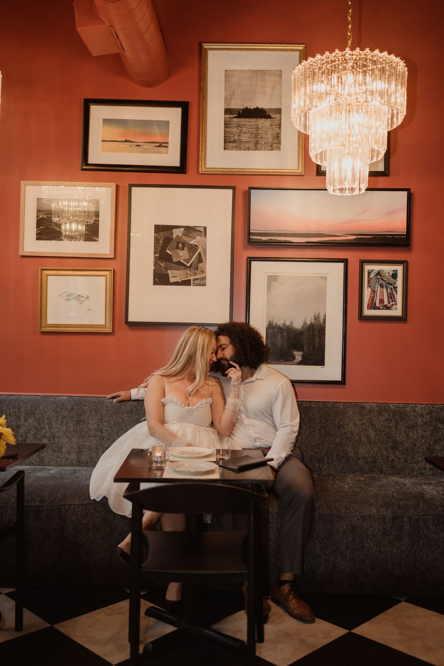 A couple sits close together on a bench beneath framed artwork and a crystal chandelier.