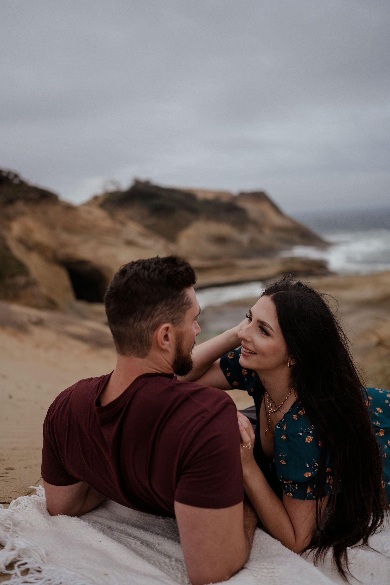 Romantic couple lying on beach blanket near coastal cliffs during overcast day.
